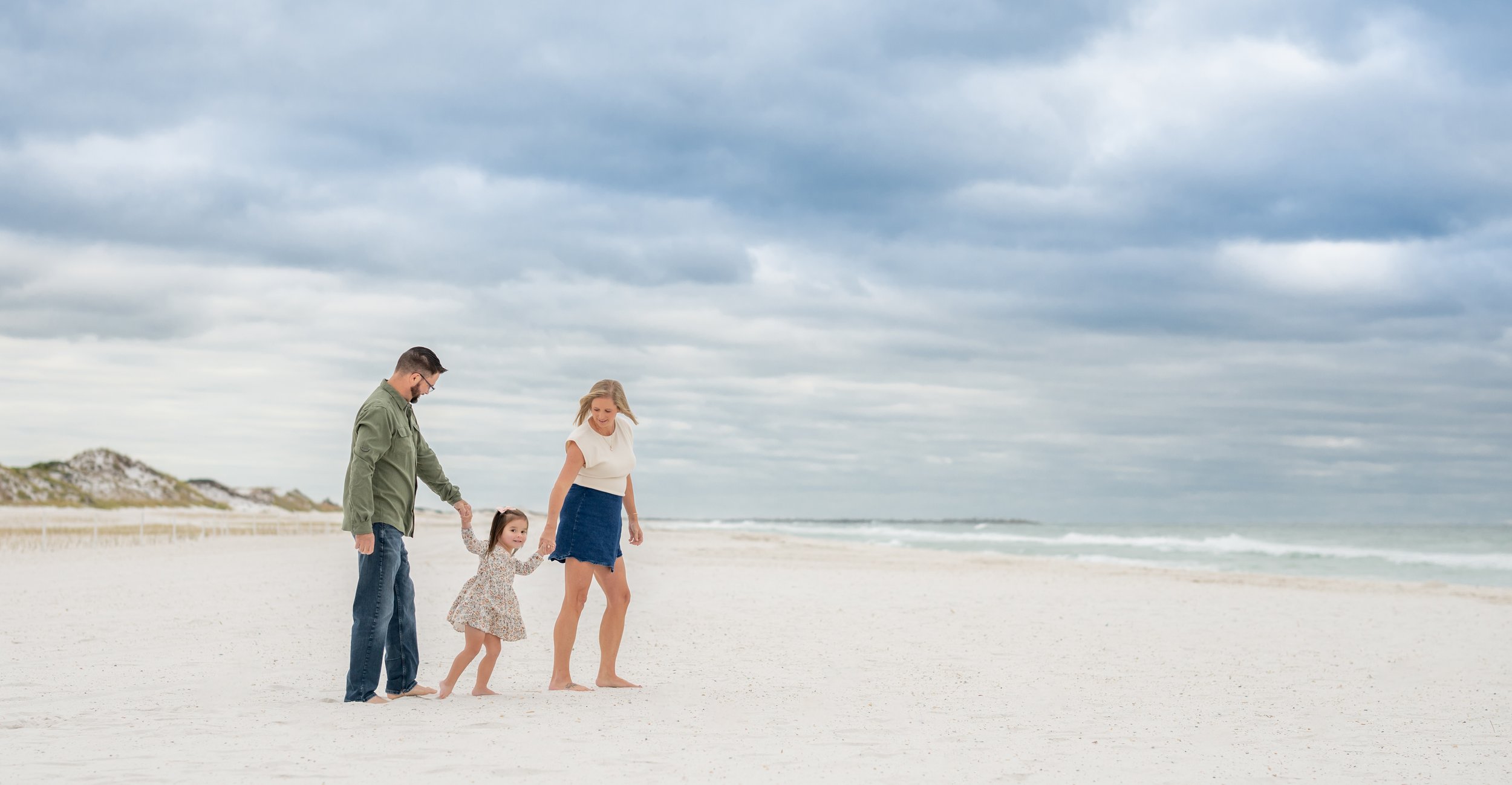 A family of three walking hand in hand on a sandy beach under a cloudy sky.