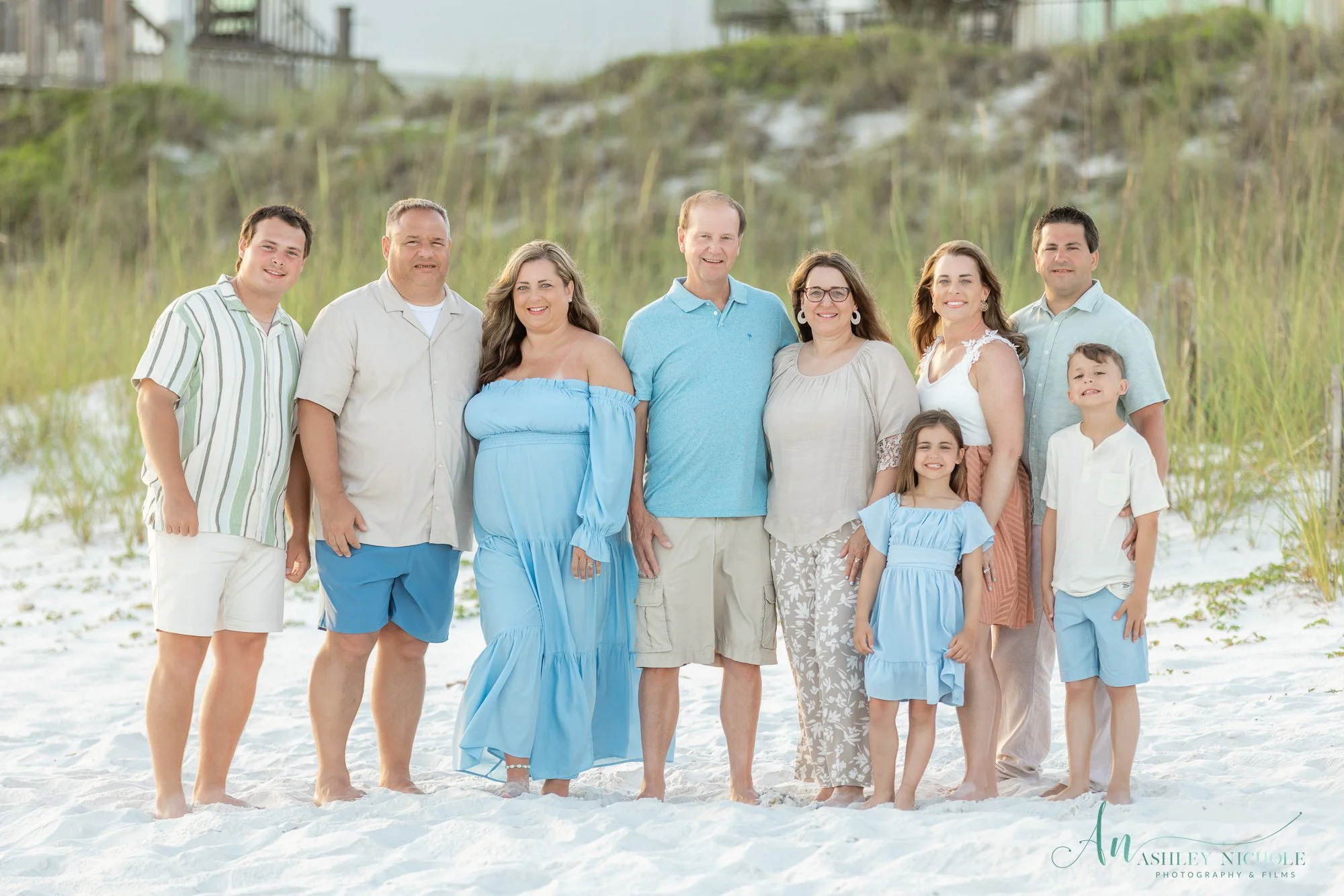 Family group standing on a sandy beach with grassy dunes in the background. There are five adults and four children, all dressed in light pastel and white clothing, smiling at the camera.