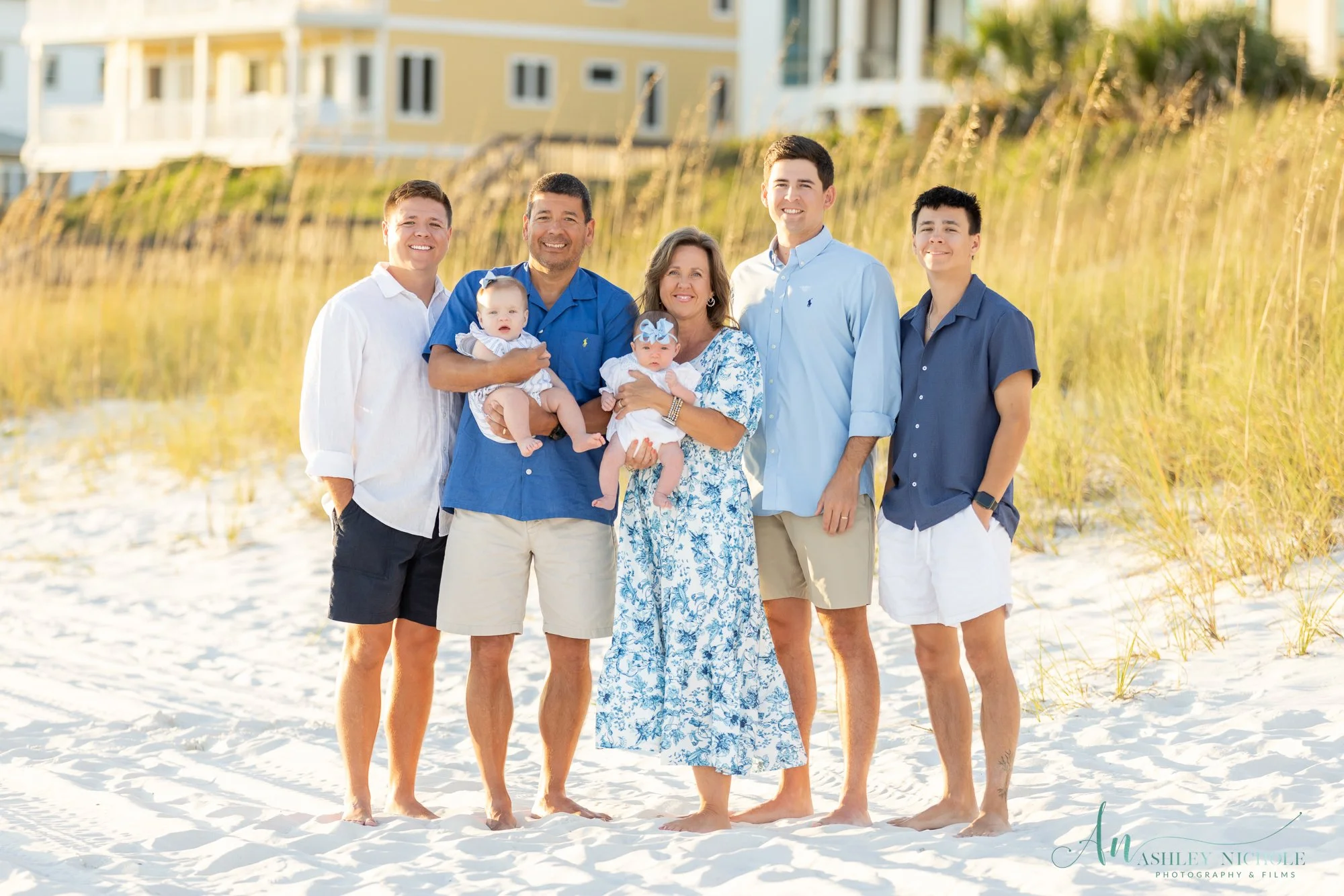 Family of seven on the beach, three adults, two teenagers, two infants, in white and blue clothing, with houses and tall grass in the background.