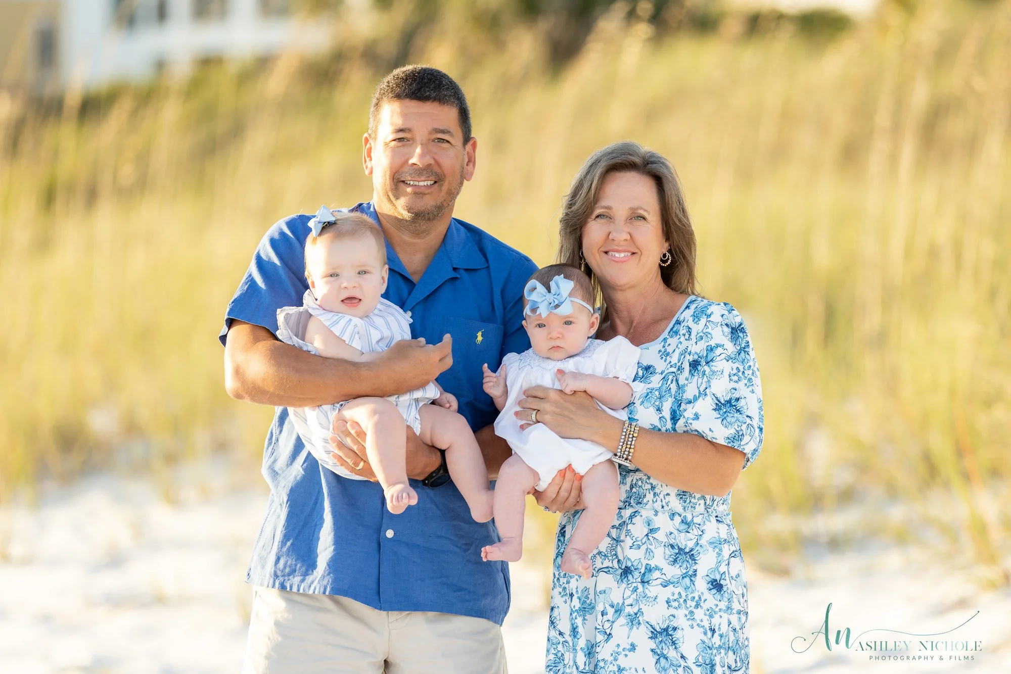 Family photo of a man, woman, and two babies outdoors on a sunny day, with tall grasses in the background. The man is wearing a blue shirt and holding one baby girl, while the woman, dressed in a floral dress, holds the other baby girl. Both babies a