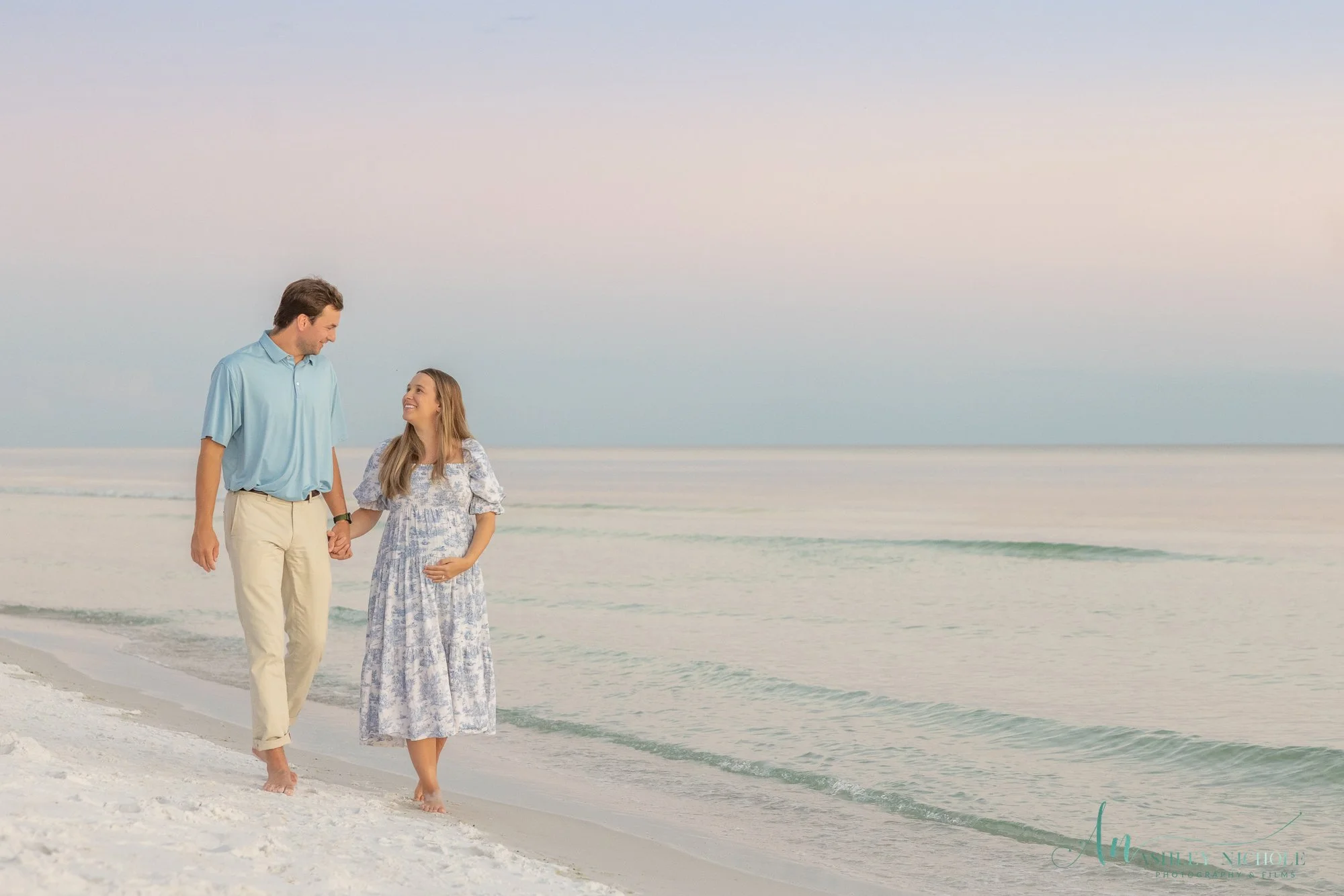 A couple holding hands and walking along the beach, smiling at each other with the ocean and sky in the background at sunset or sunrise.