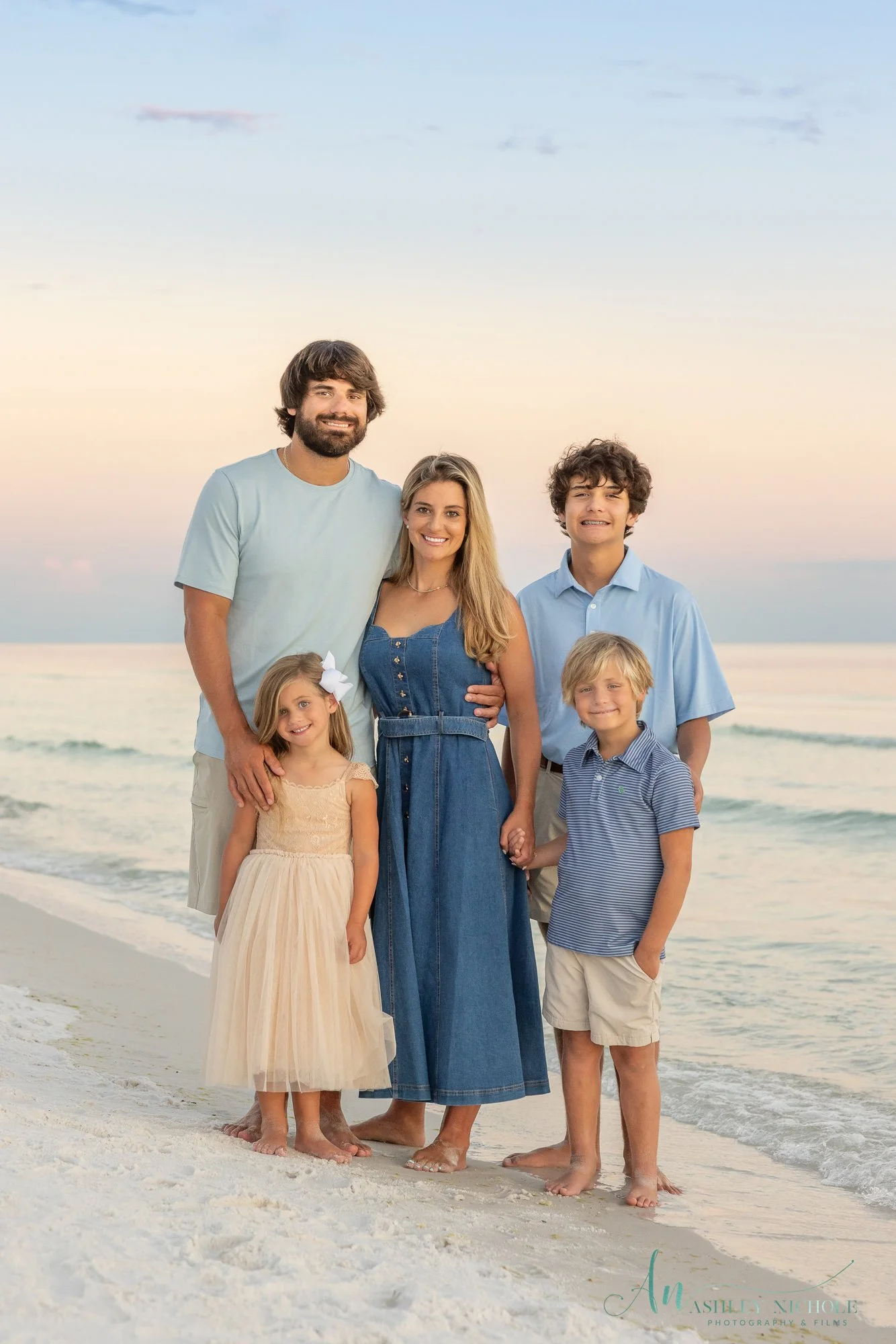 Family of six standing on the beach at sunset, holding hands, smiling, wearing casual summer clothes.