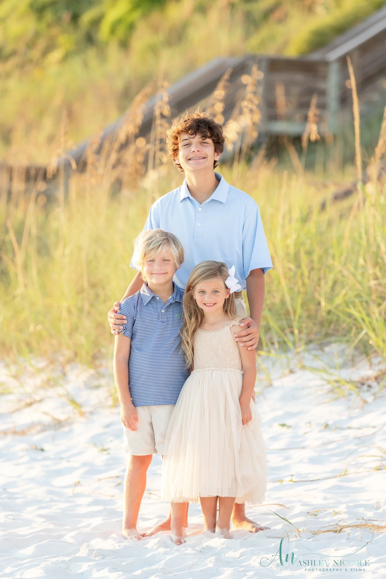 A family of four standing close together on a sandy beach with tall grasses and a weathered wooden structure in the background, smiling at the camera.