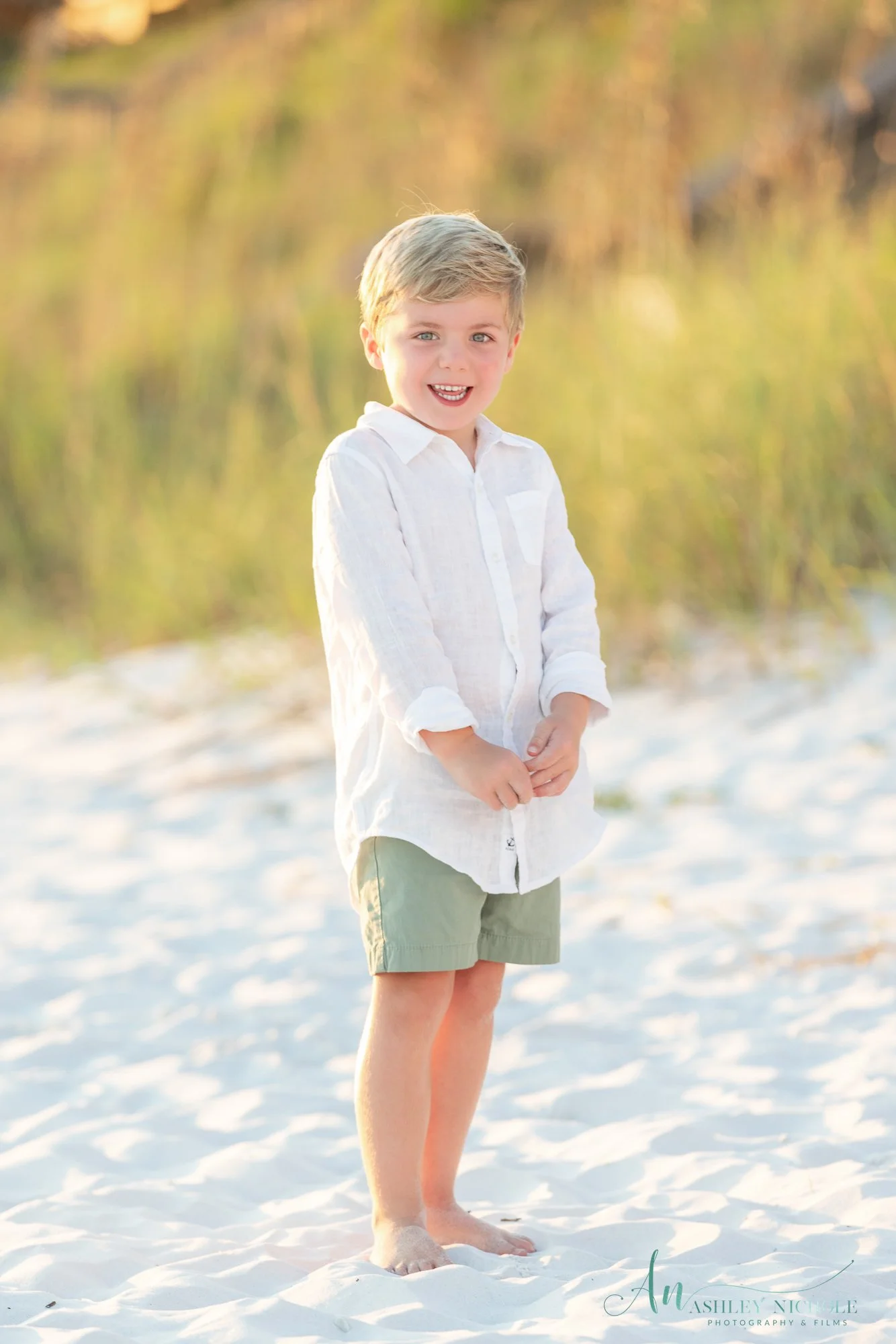A young boy with blonde hair, wearing a white shirt and light green shorts, standing barefoot on white sand at the beach with trees and greenery in the background, smiling and looking at the camera during sunset.