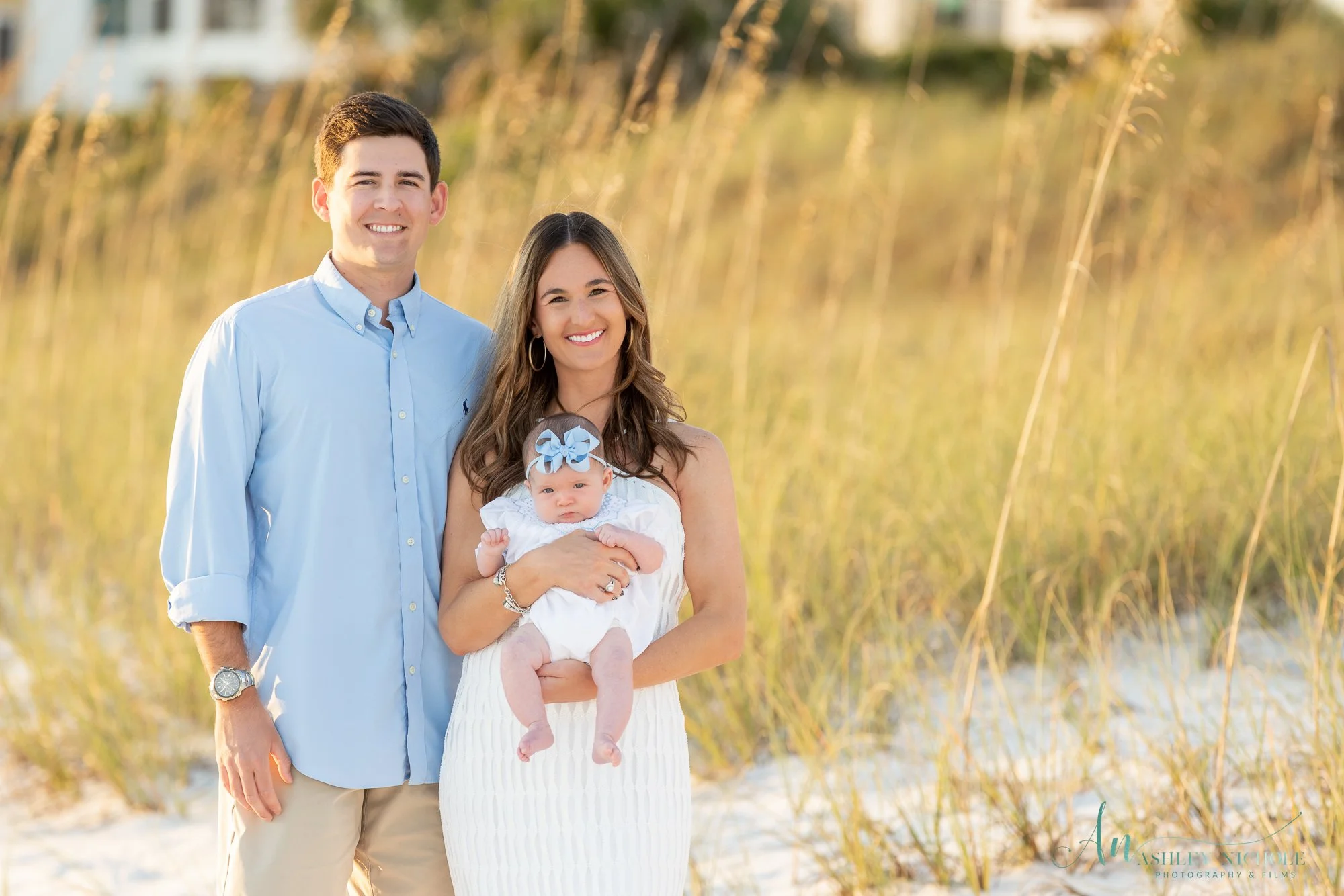 A smiling family of three standing outdoors at the beach, with a man in a light blue shirt, a woman in a white dress holding a baby girl with a blue bow headband, and tall grass in the background.