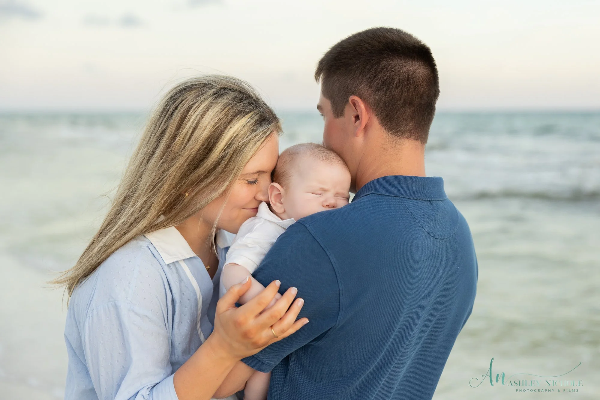 A family of three, mother, father, and baby, sharing a warm moment on the beach.