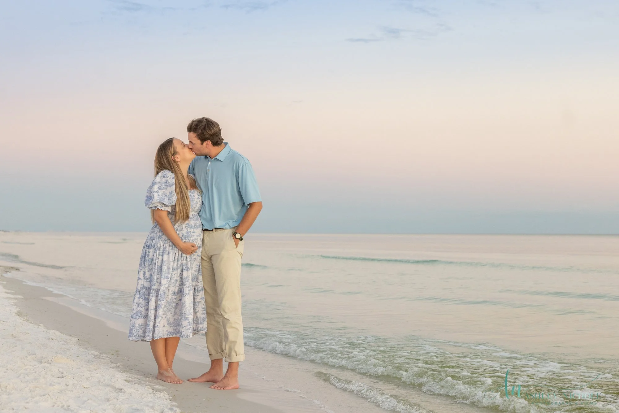 A romantic couple kissing on the beach at sunset, standing barefoot near the shoreline with gentle waves and pastel-colored sky.