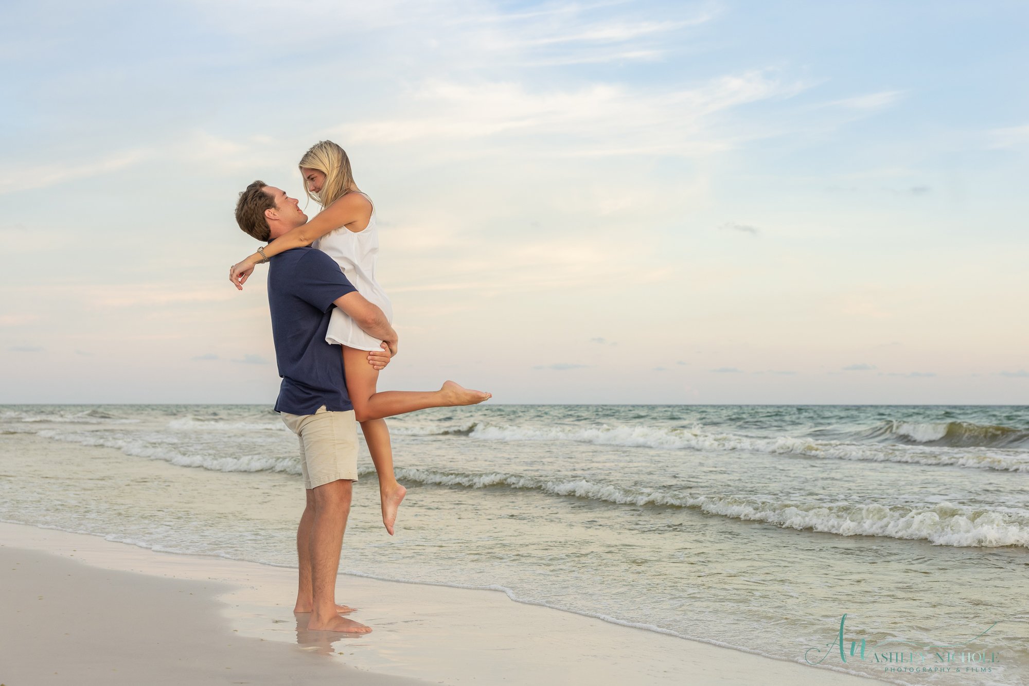 A man holding a woman in his arms on a beach with the ocean in the background.