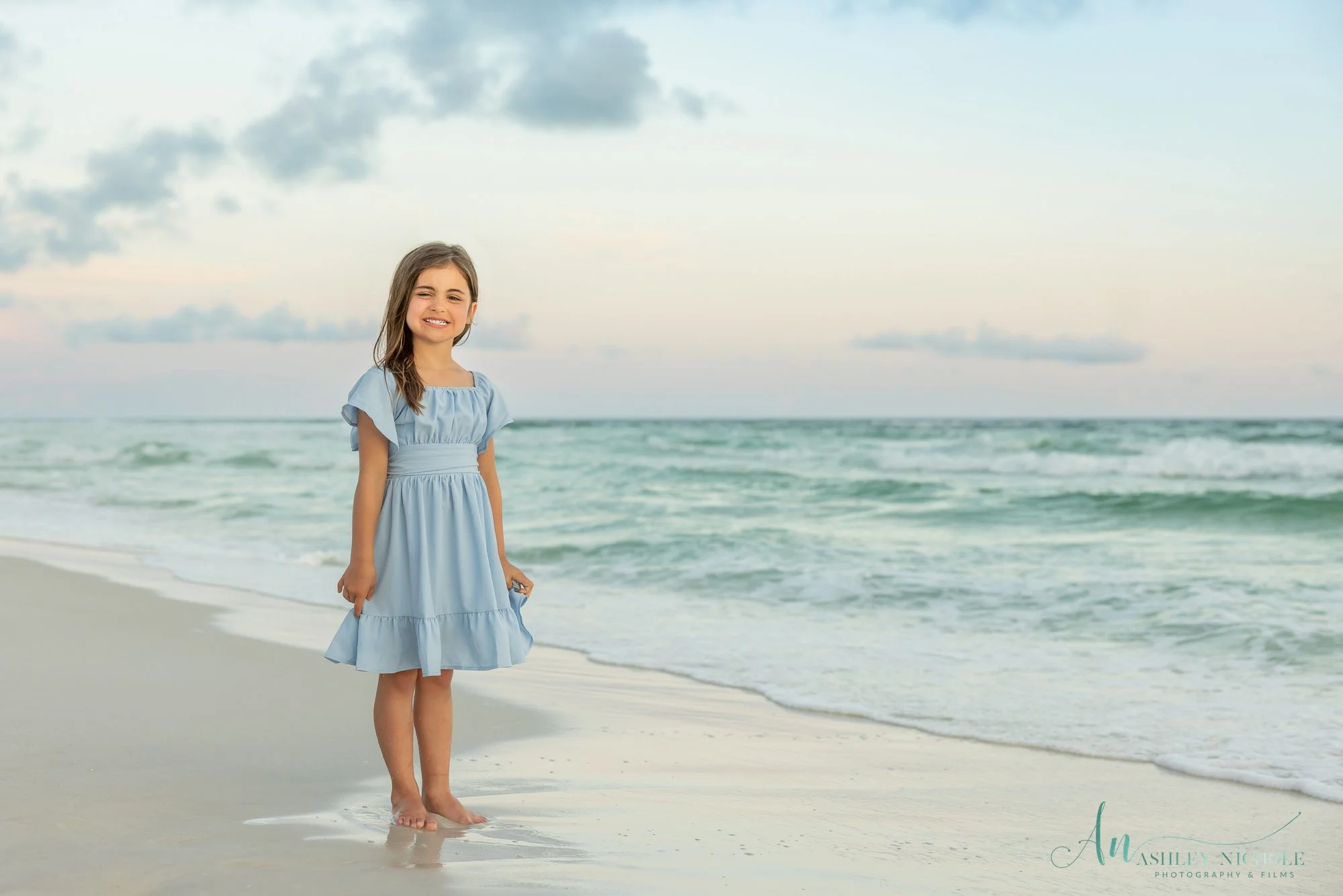 A young girl with long brown hair wearing a light blue dress standing barefoot on a sandy beach near the shoreline with calm ocean waves in the background and a cloudy sky.