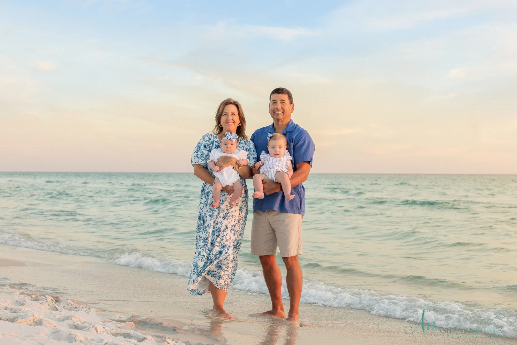 A family of four standing on a beach at sunset, smiling. The mother and father each hold a baby; the mother wears a long floral dress, and the father maintains a casual blue shirt and khakis. The beach features white sand and calm ocean waves in the 