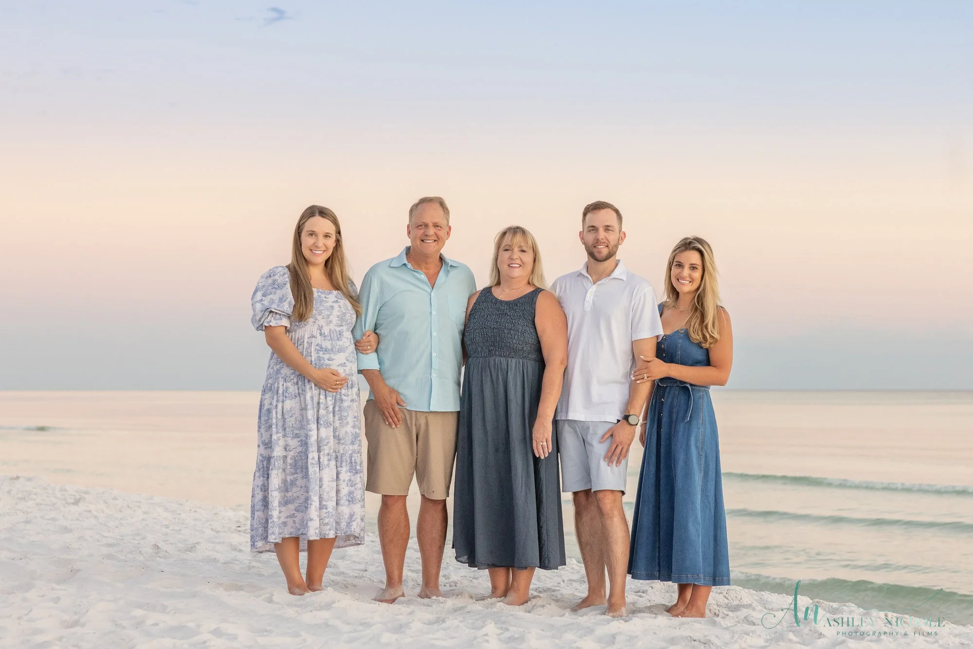 A family of six standing on a beach at sunset, smiling, with the ocean in the background.