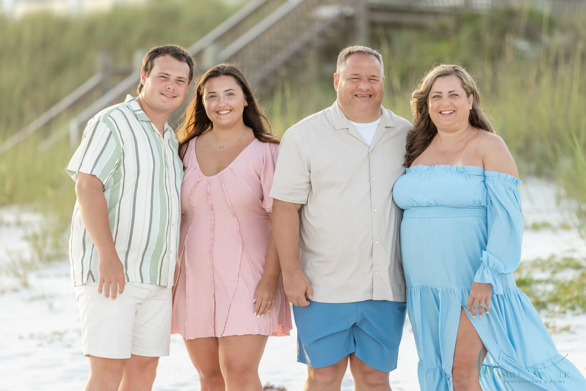 A family of five standing on a beach, smiling for a photo, with a staircase and greenery in the background.