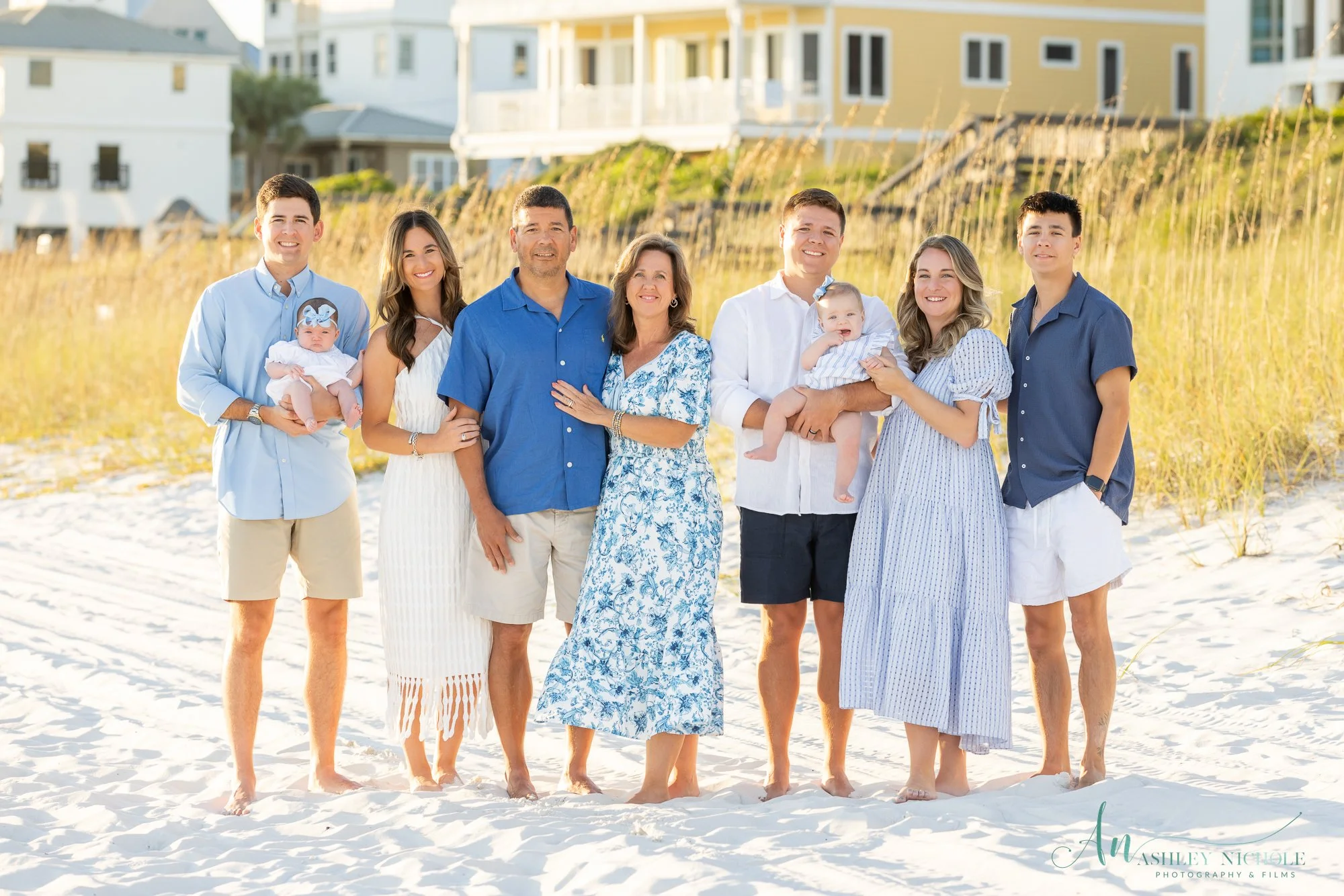 A large family stands on a sandy beach with tall grass and coastal houses in the background, all dressed in summer clothing, smiling at the camera.