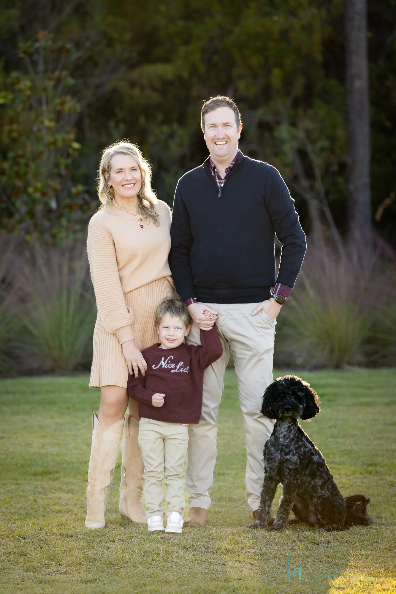 Family of four with a woman, a man, a boy labeled Nick List, and a black curly-haired dog outdoors on a grassy field with trees and bushes in the background, smiling at the camera.