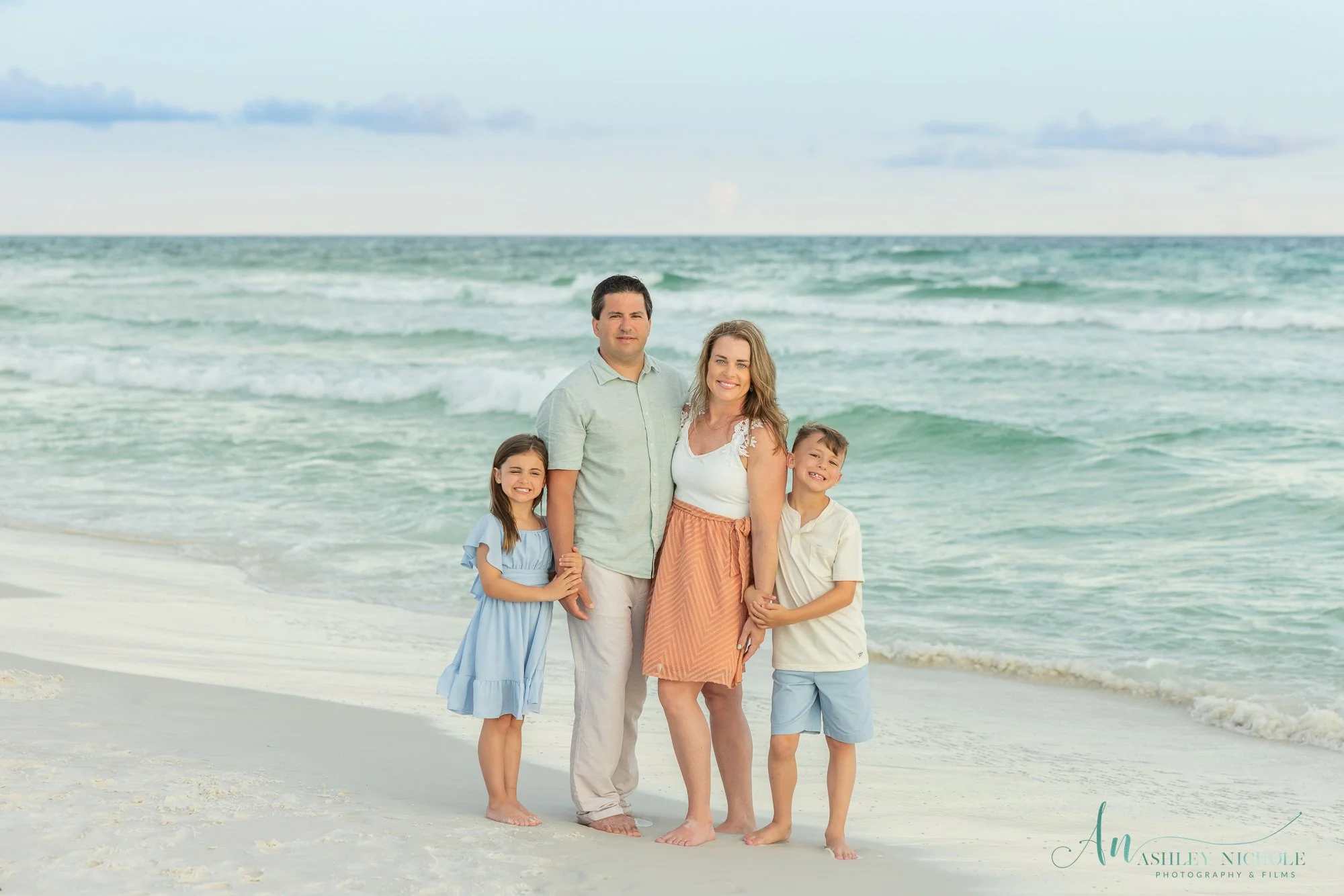 A family of four standing on a sandy beach by the ocean, smiling and holding hands. The family includes a woman, a man, a young girl in a blue dress, and a young boy in a white shirt and blue shorts. The ocean waves are in the background under a part
