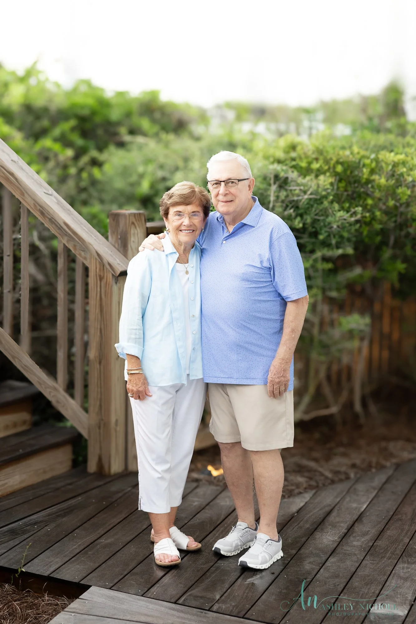 An elderly couple standing on a wooden deck outdoors, smiling and hugging each other, with greenery in the background.