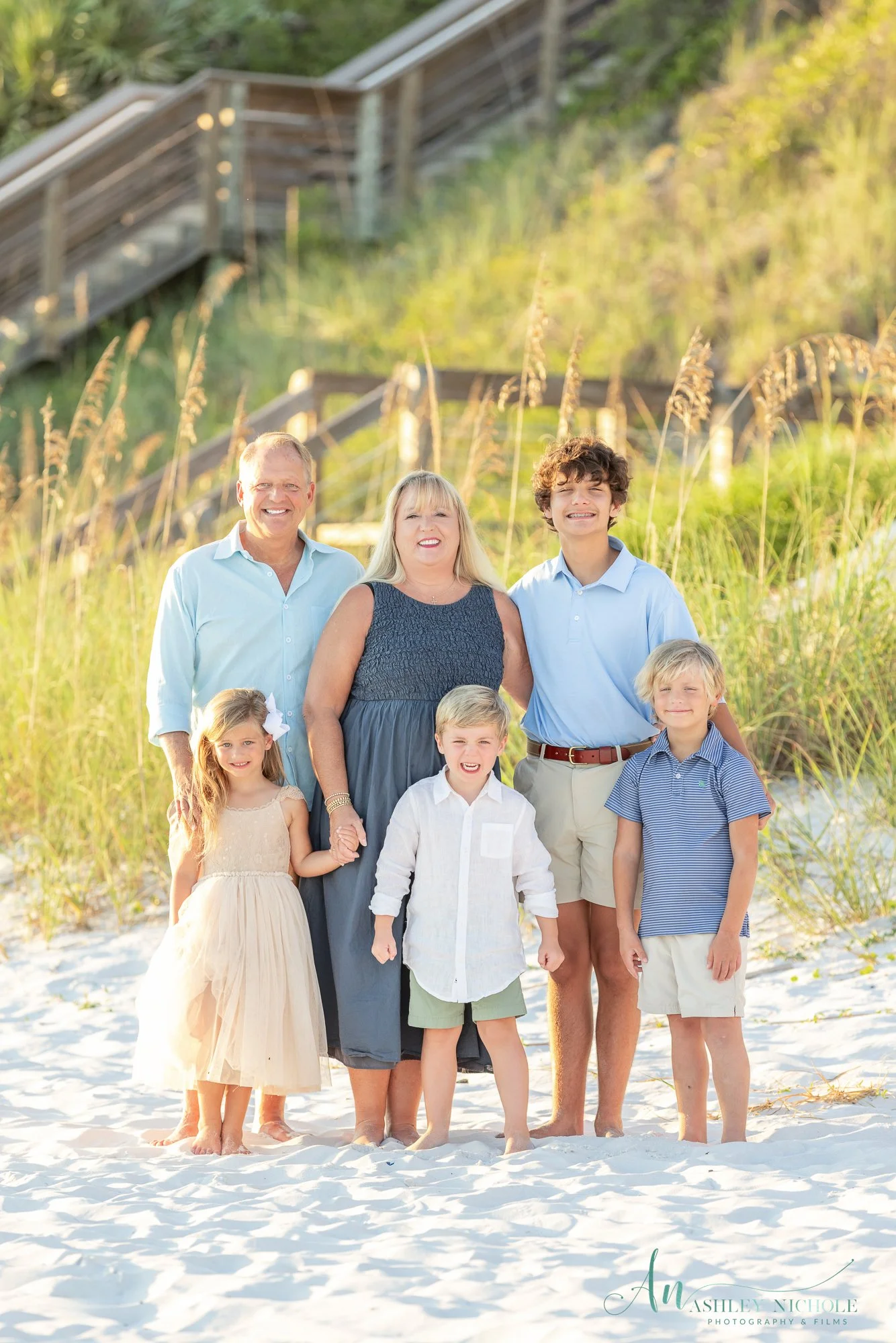 Family of seven standing on a sandy beach, smiling with grass and stairs in the background.