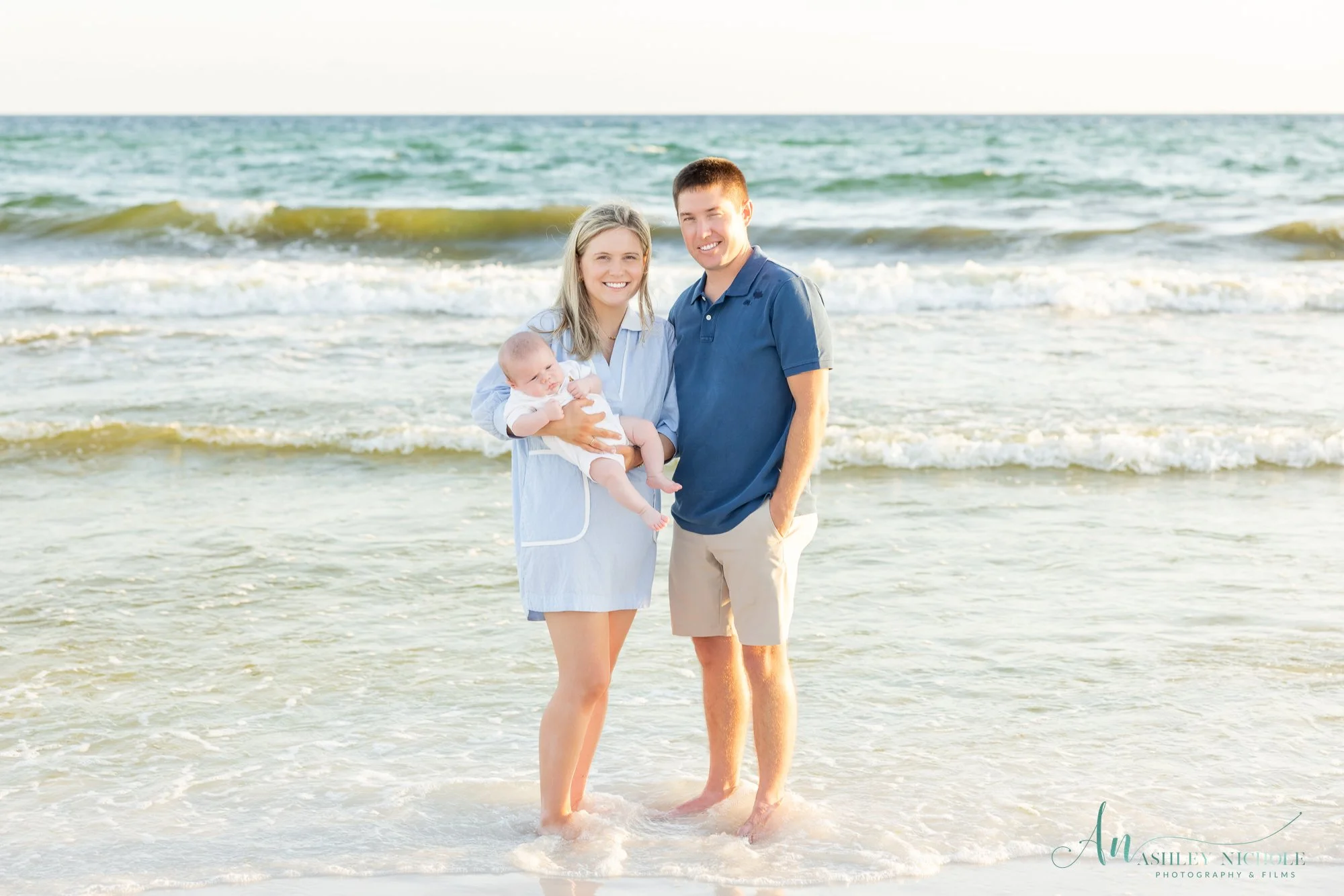 Family of three standing in the ocean waves at the beach during sunset, woman holding a baby boy, man standing next to them, smiling.