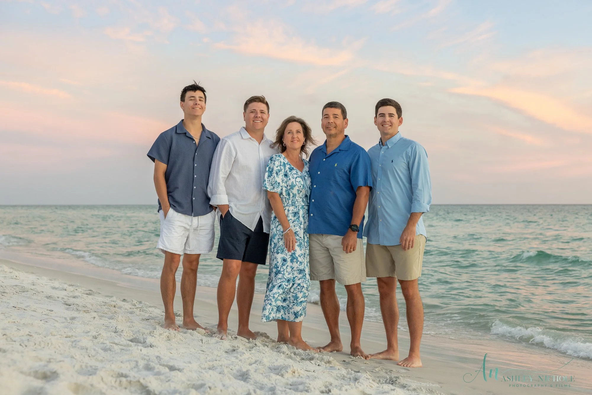 Family of six standing on the beach near the ocean with a sunset sky in the background. They are smiling and dressed in casual summer clothing.