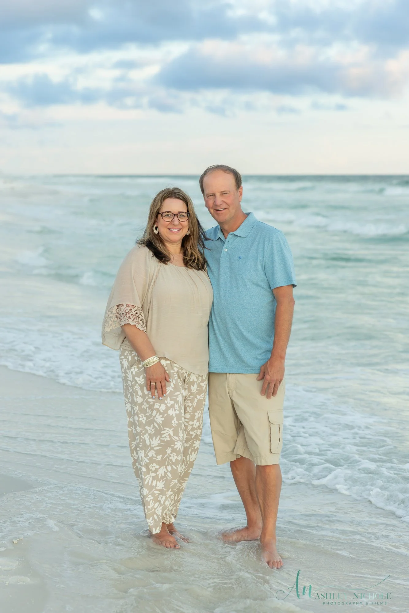 A couple standing on the beach at the shoreline with the ocean and cloudy sky in the background, both smiling and dressed in casual summer clothing.