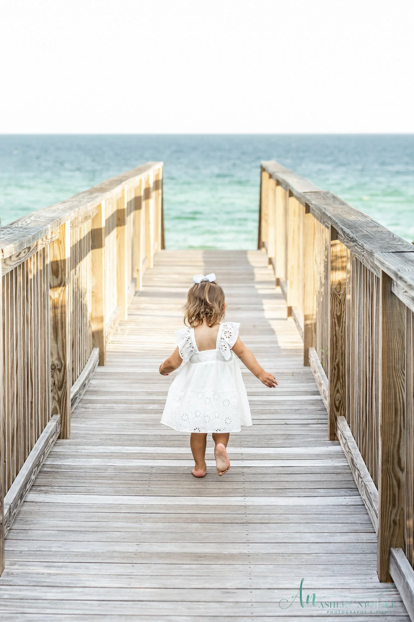 A young girl in a white dress with a white bow on her head walking barefoot on a wooden pier towards the ocean.