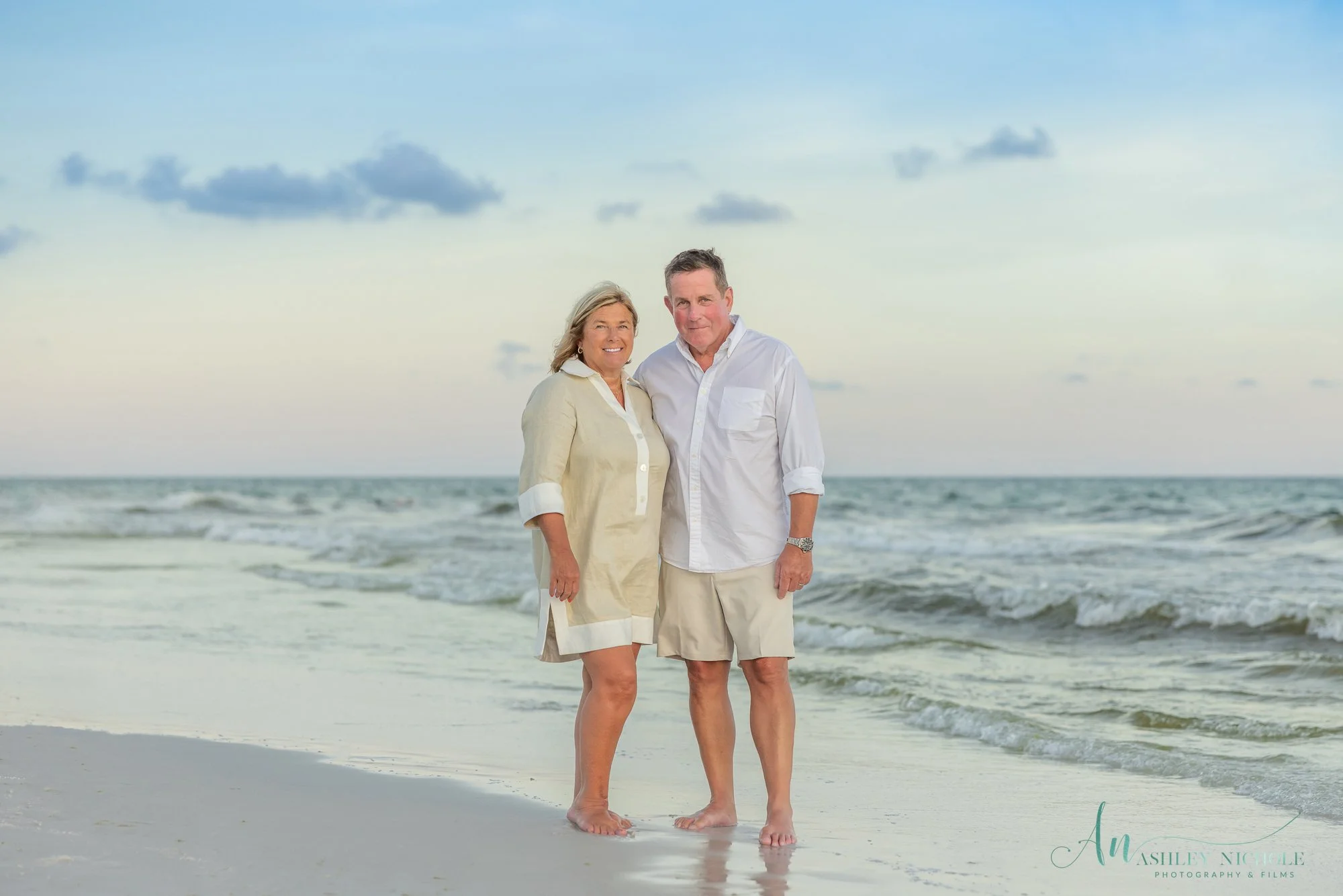 A middle-aged couple standing barefoot on the beach at sunset, smiling at the camera with the ocean and sky in the background.