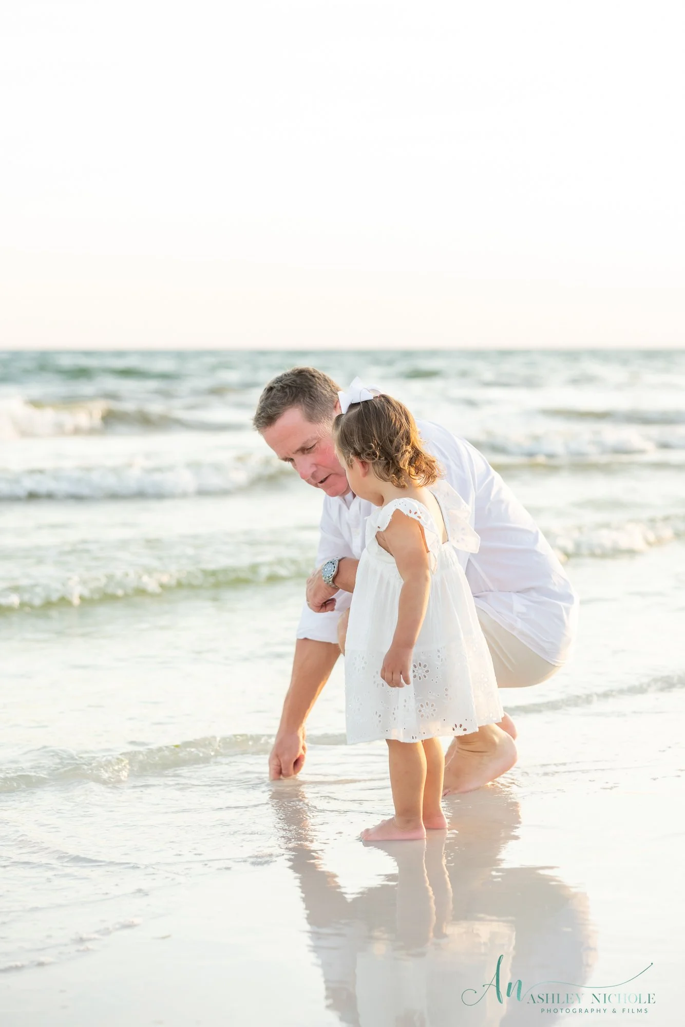 A man and a young girl, both dressed in white, are on a beach, kneeling by the water, looking at the sand and waves.