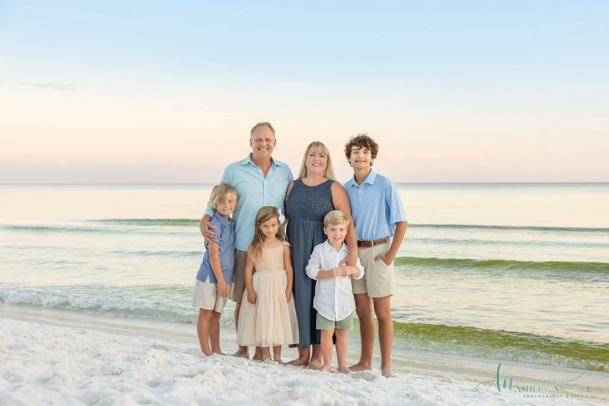 Family of seven standing on a beach with ocean waves in the background, smiling at the camera during sunset or sunrise.