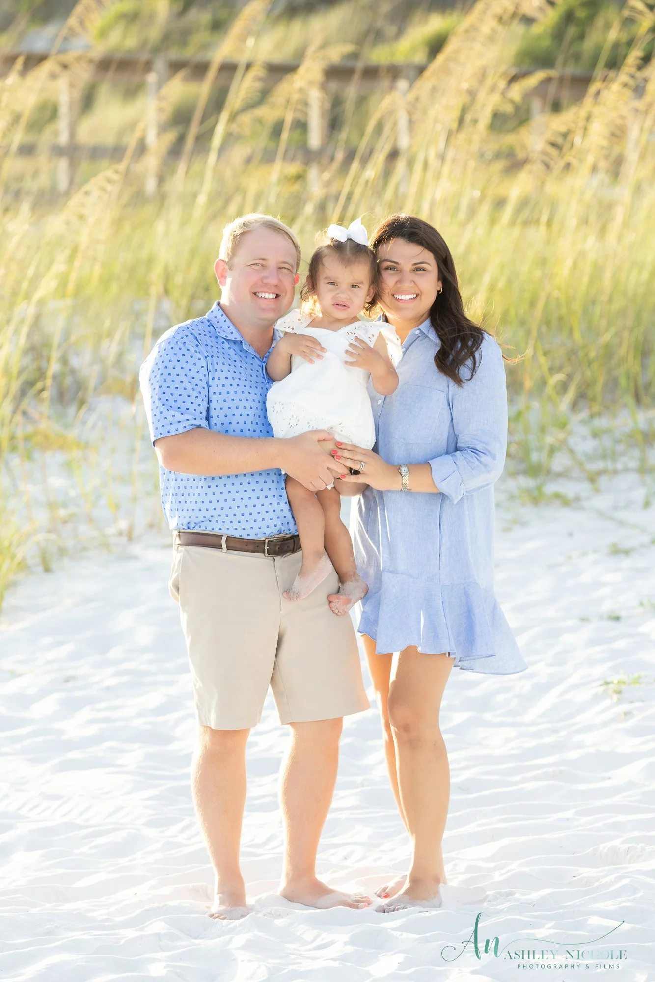 Family of three at the beach, smiling, with a man holding a young girl in a white dress and a woman in a blue dress, standing on white sand with grass in the background.