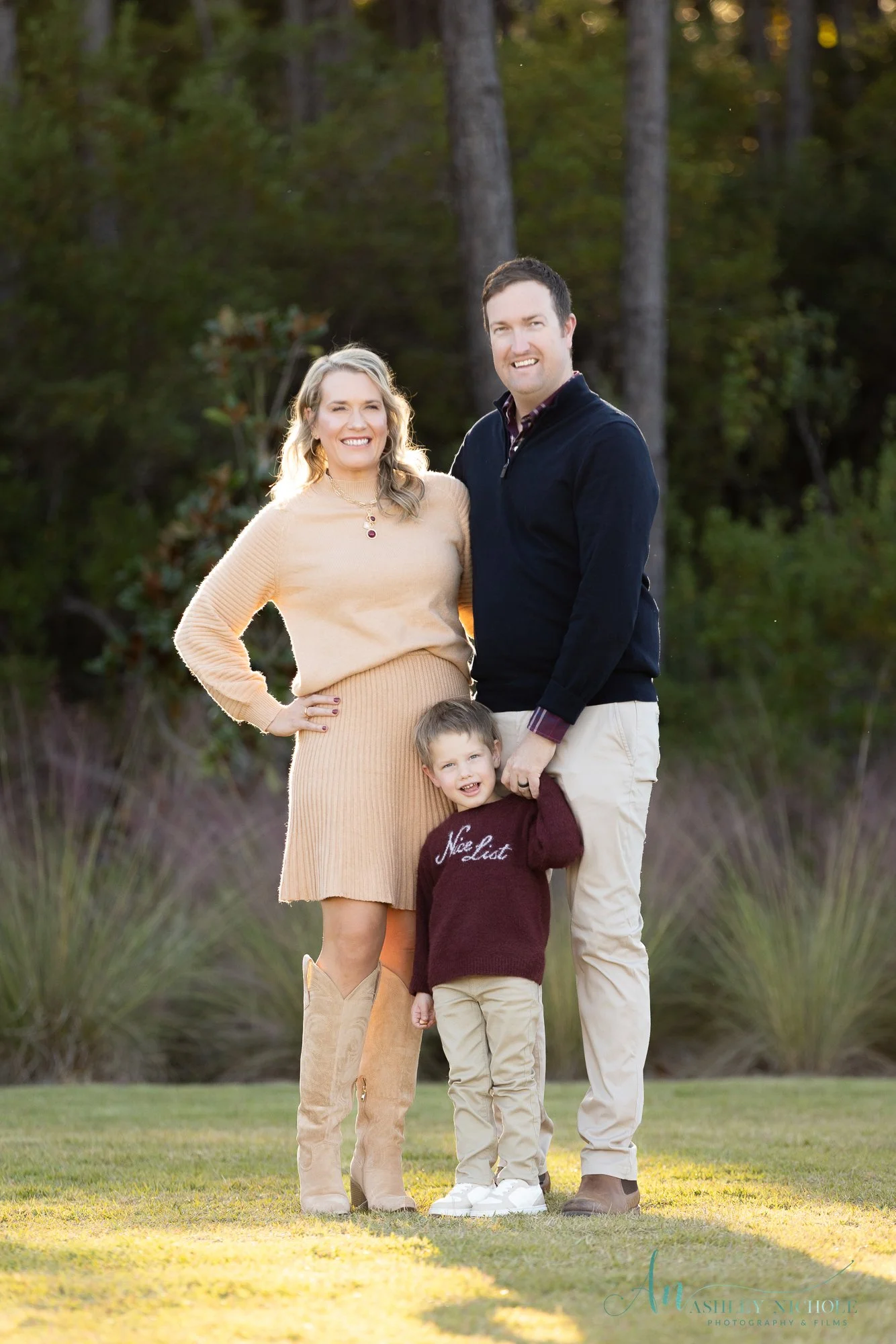 A family of three standing outdoors on the grass, smiling at the camera. The woman is wearing a beige sweater and skirt, the man is wearing a black jacket and beige pants, and the young boy is wearing a burgundy sweater with 'Nice List' written on it