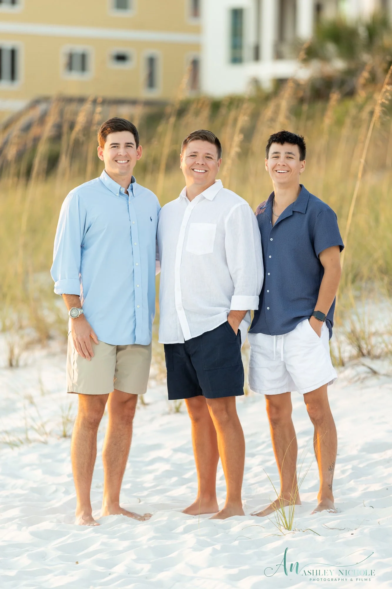 Three young men standing on a sandy beach with grass and a building in the background, smiling at the camera during sunset.