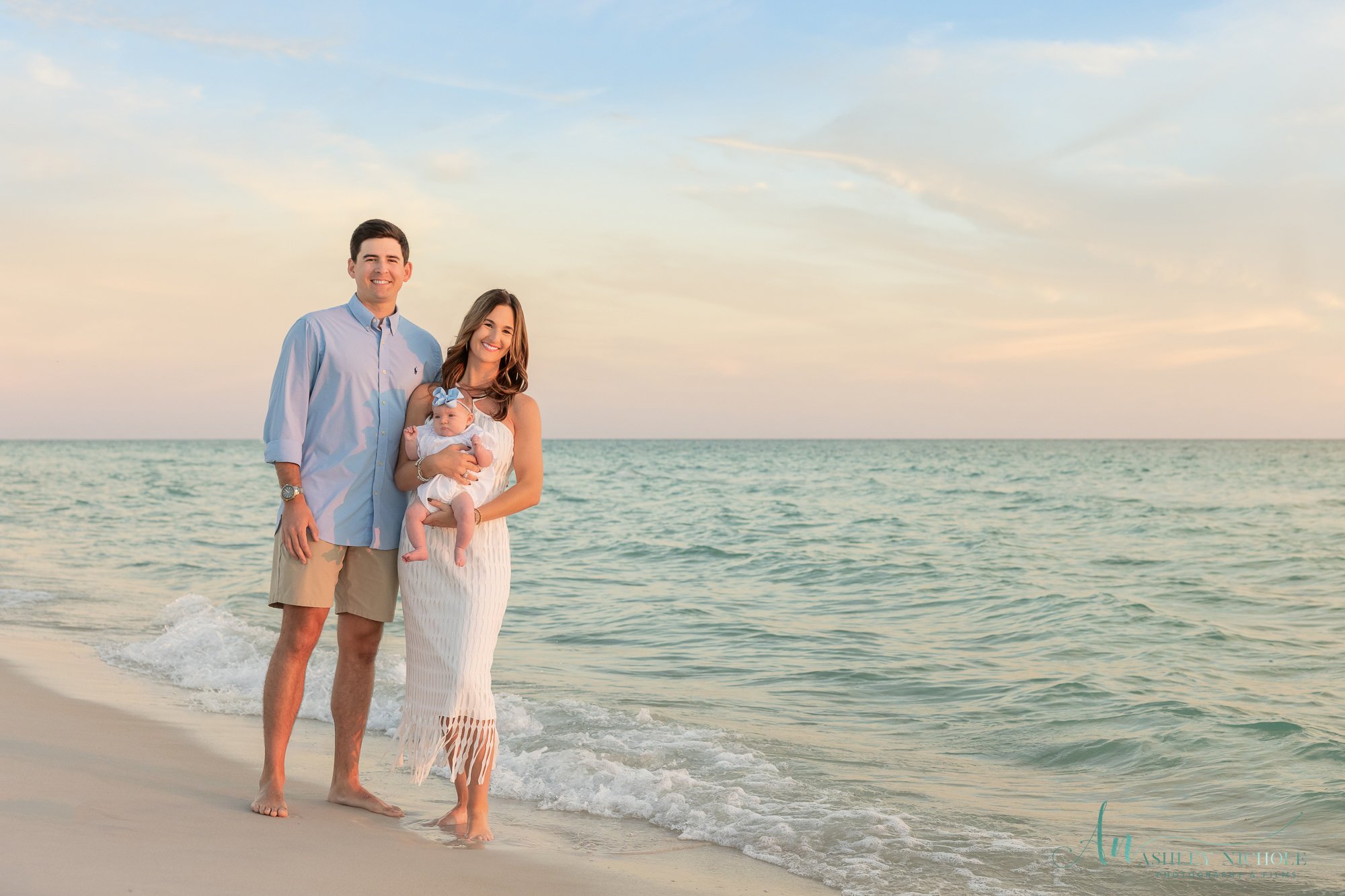 A family of three standing on the beach near the ocean at sunset, with a man, woman, and baby, all smiling and looking at the camera.
