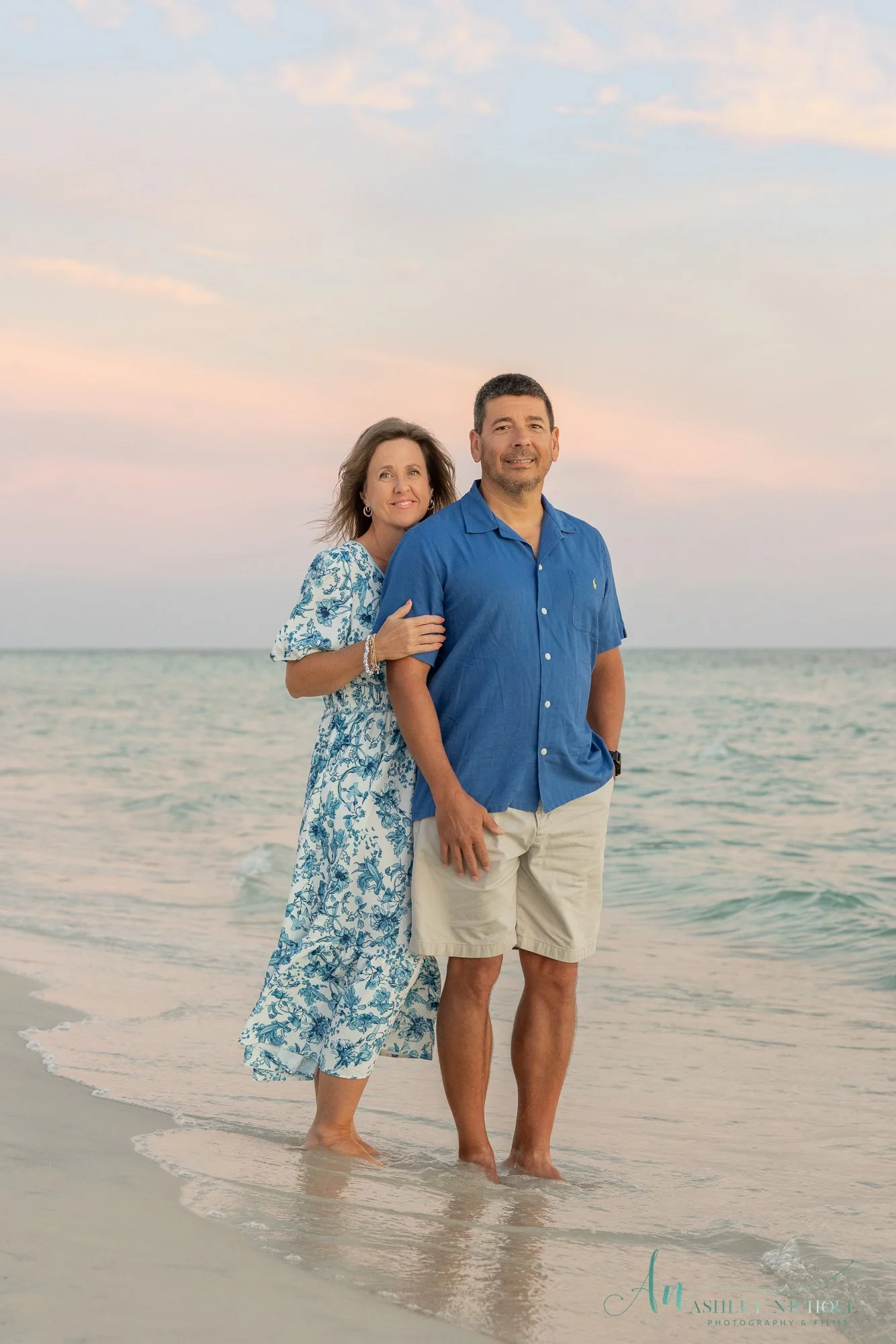 A couple standing in shallow ocean water at sunset, with the woman leaning on the man's shoulder and both smiling at the camera.