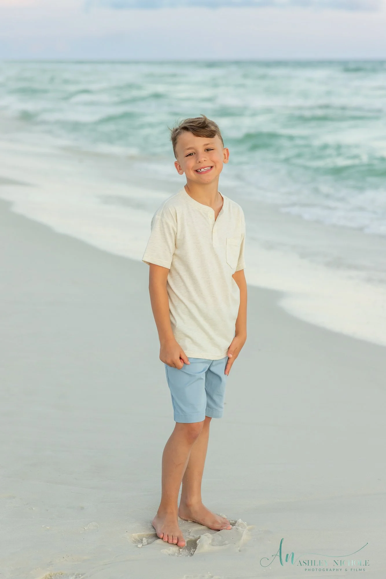 A smiling boy standing on the beach with ocean waves behind him, wearing a light-colored t-shirt and light blue shorts, barefoot on the sand.