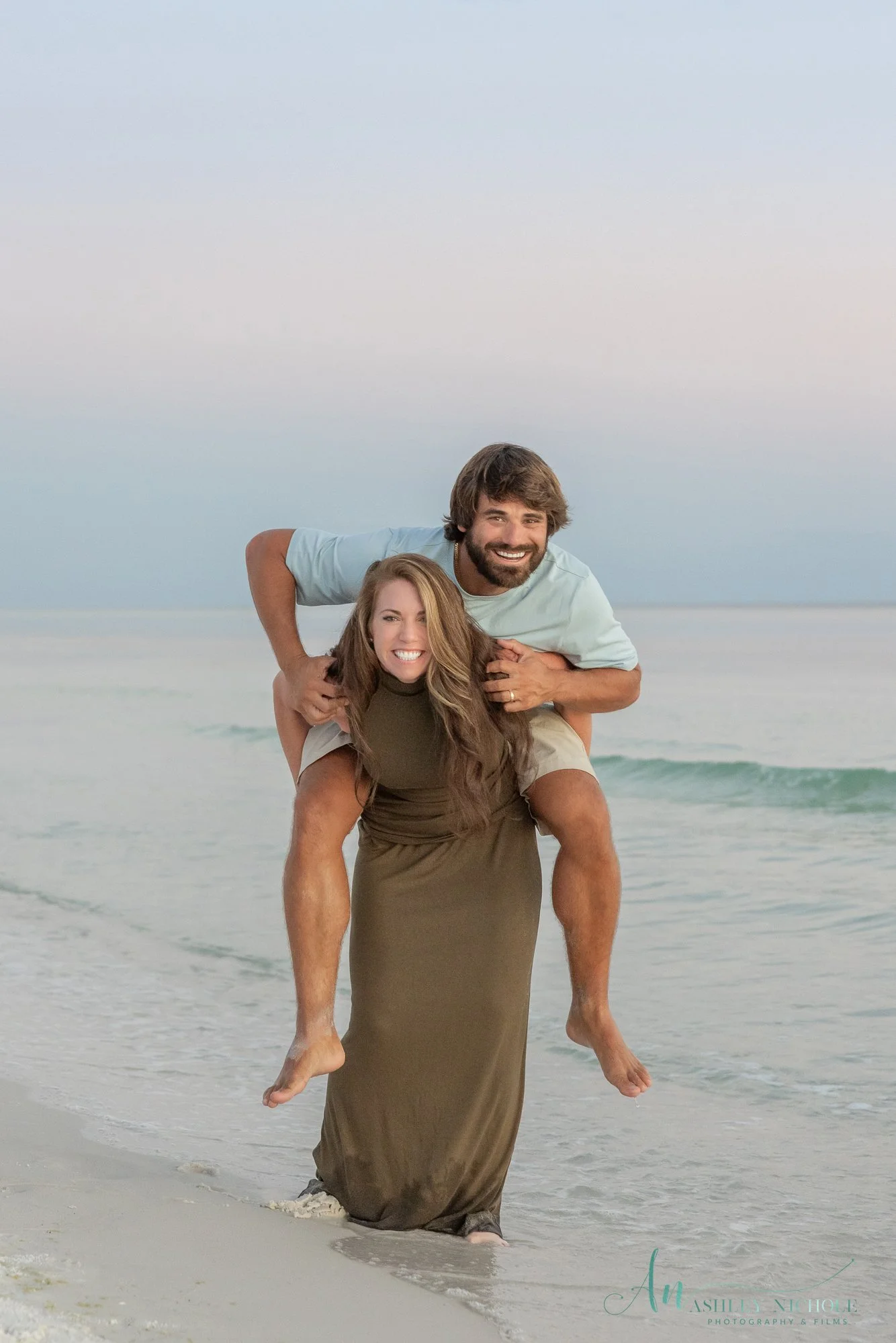 A smiling man giving a piggyback ride to a smiling woman along the beach shoreline.