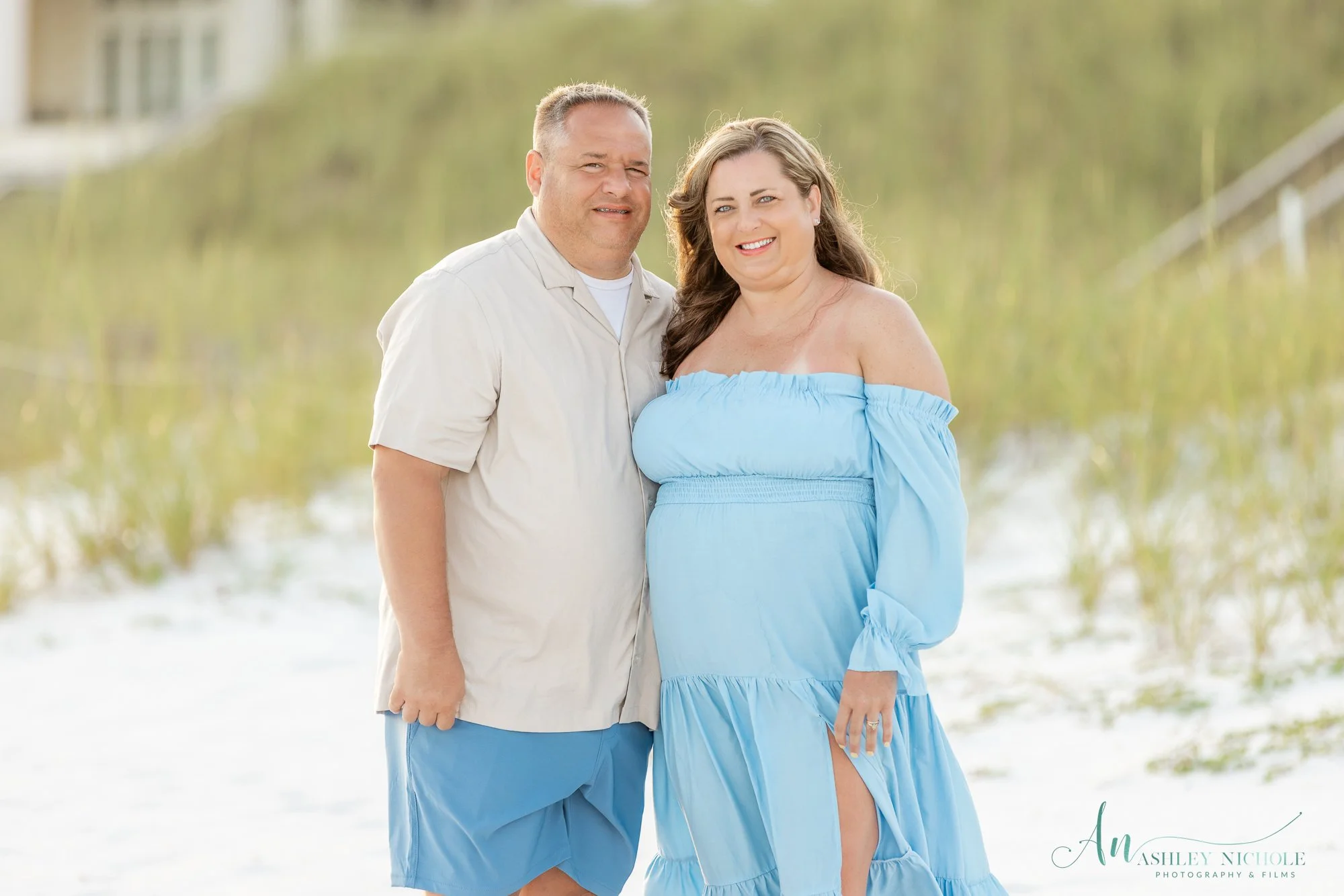A smiling pregnant woman in a blue off-shoulder dress standing with a man in a white shirt and blue shorts on a beach with green dunes in the background.