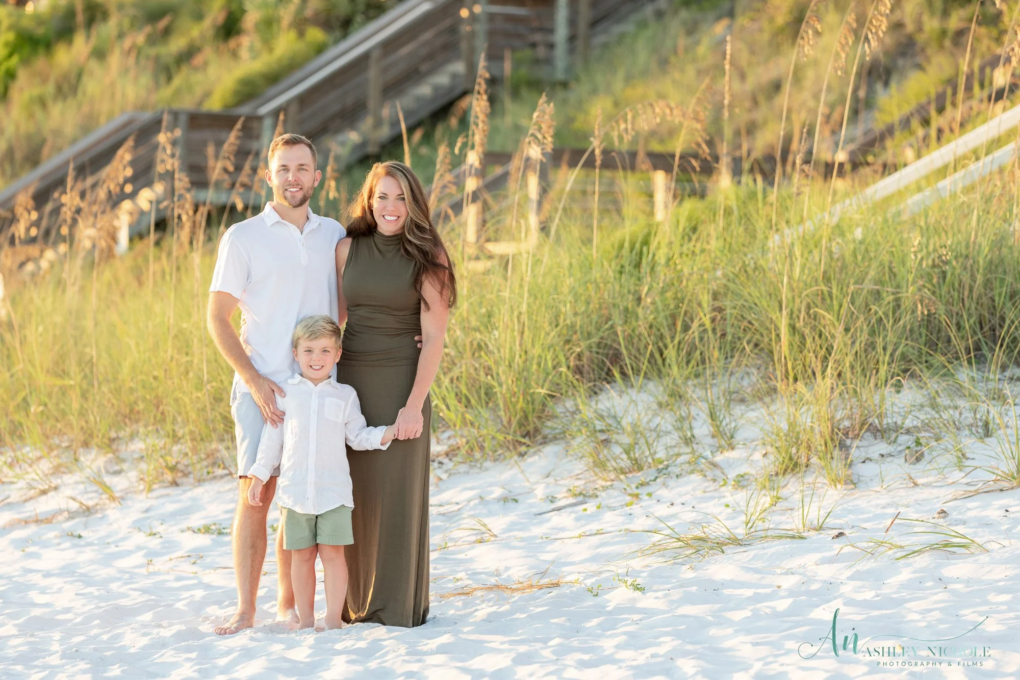 A family of three standing on a sandy beach with a wooden staircase in the background, surrounded by beach grass, during sunset.