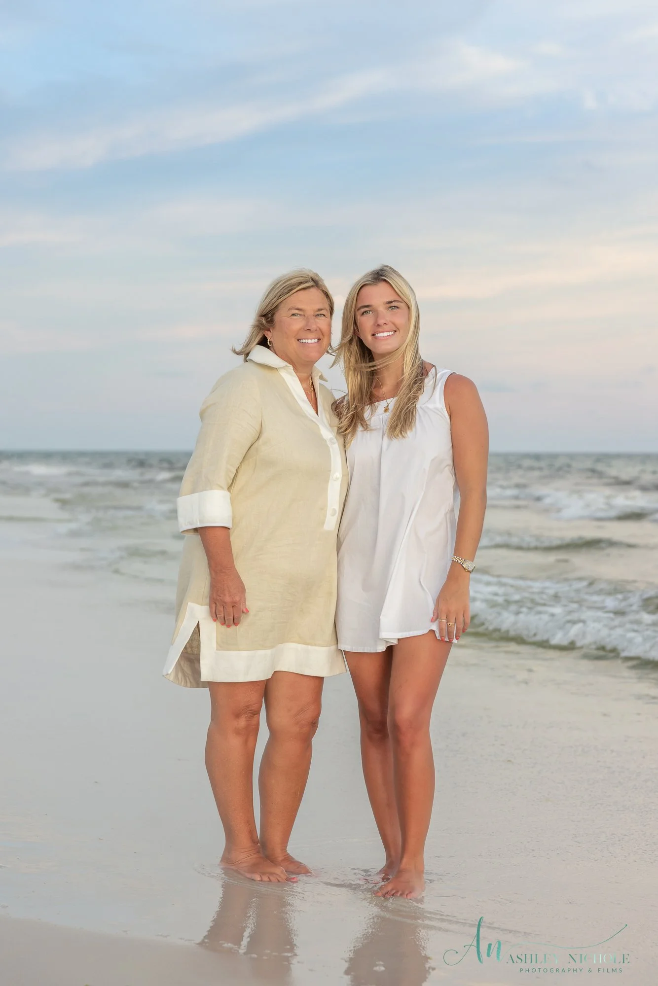 Two women stand barefoot on the beach near the water, smiling at the camera with waves in the background during sunset or sunrise.