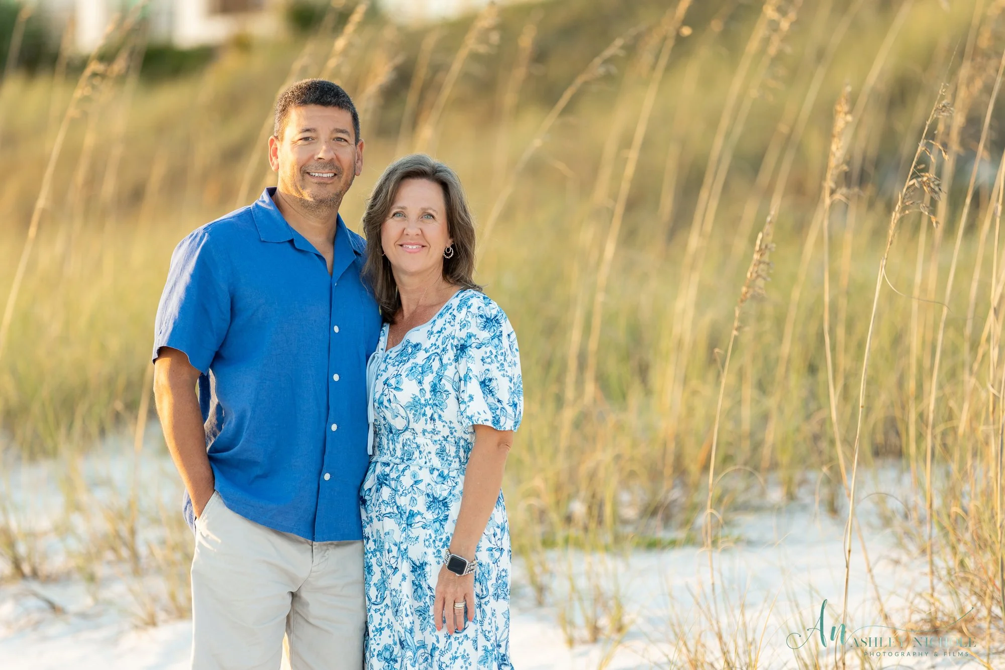 A smiling middle-aged man and woman standing close together outdoors on a sandy beach with tall grass in the background, during sunset.