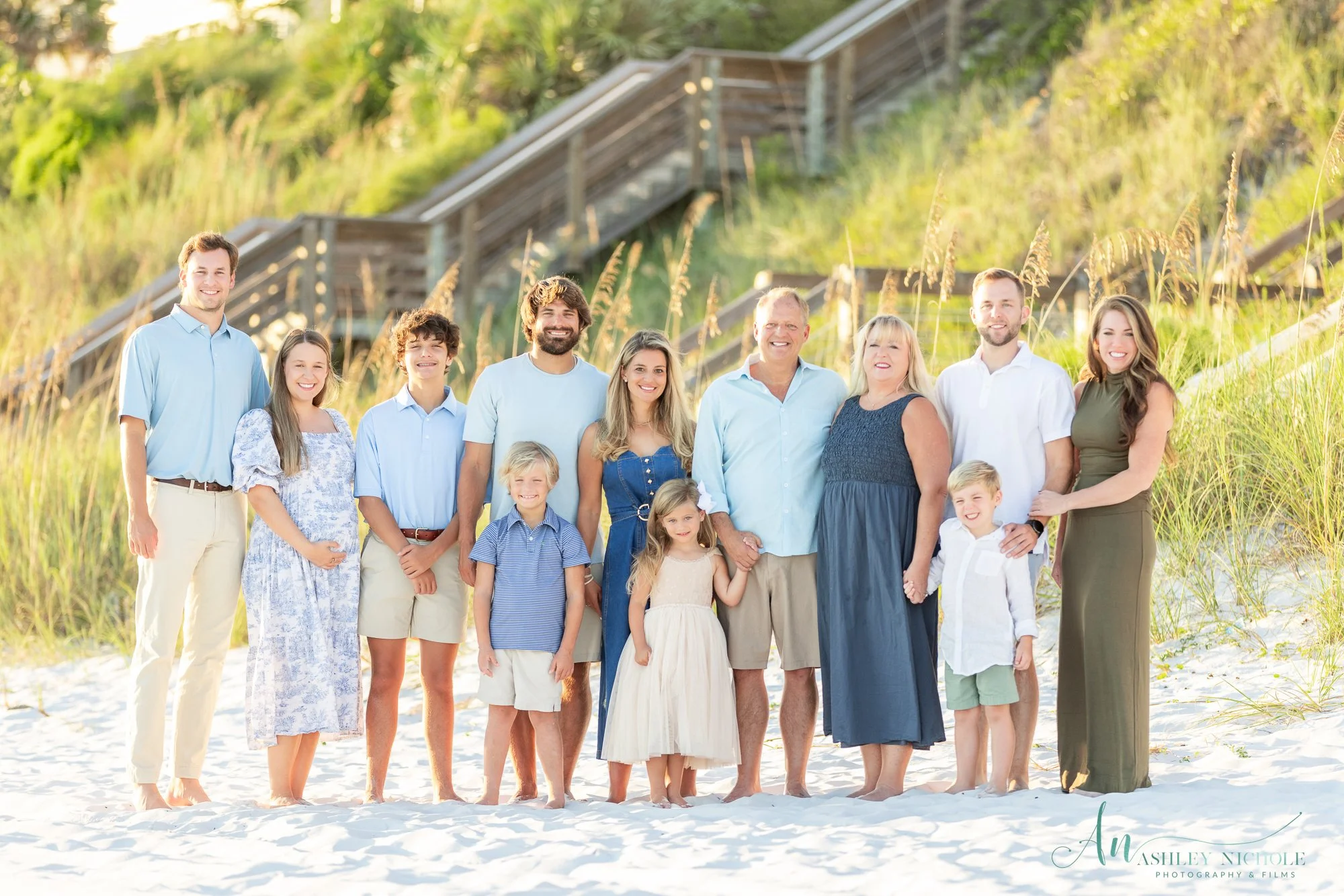 Family group standing on a sandy beach with grassy dunes and wooden stairs in the background, smiling for a photo.