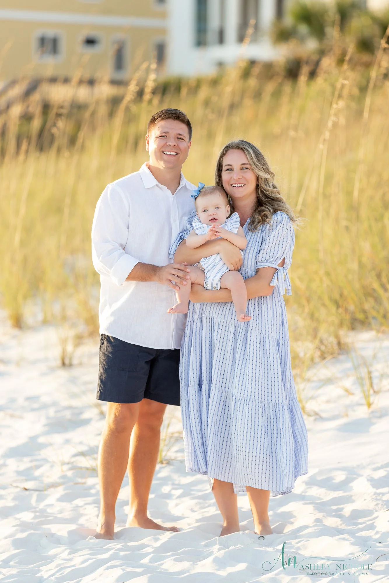A happy family of three standing on a sandy beach with grass and houses in the background. The family consists of a man, woman, and a young girl being held by the woman. They are dressed in light, summer clothing and are smiling at the camera.