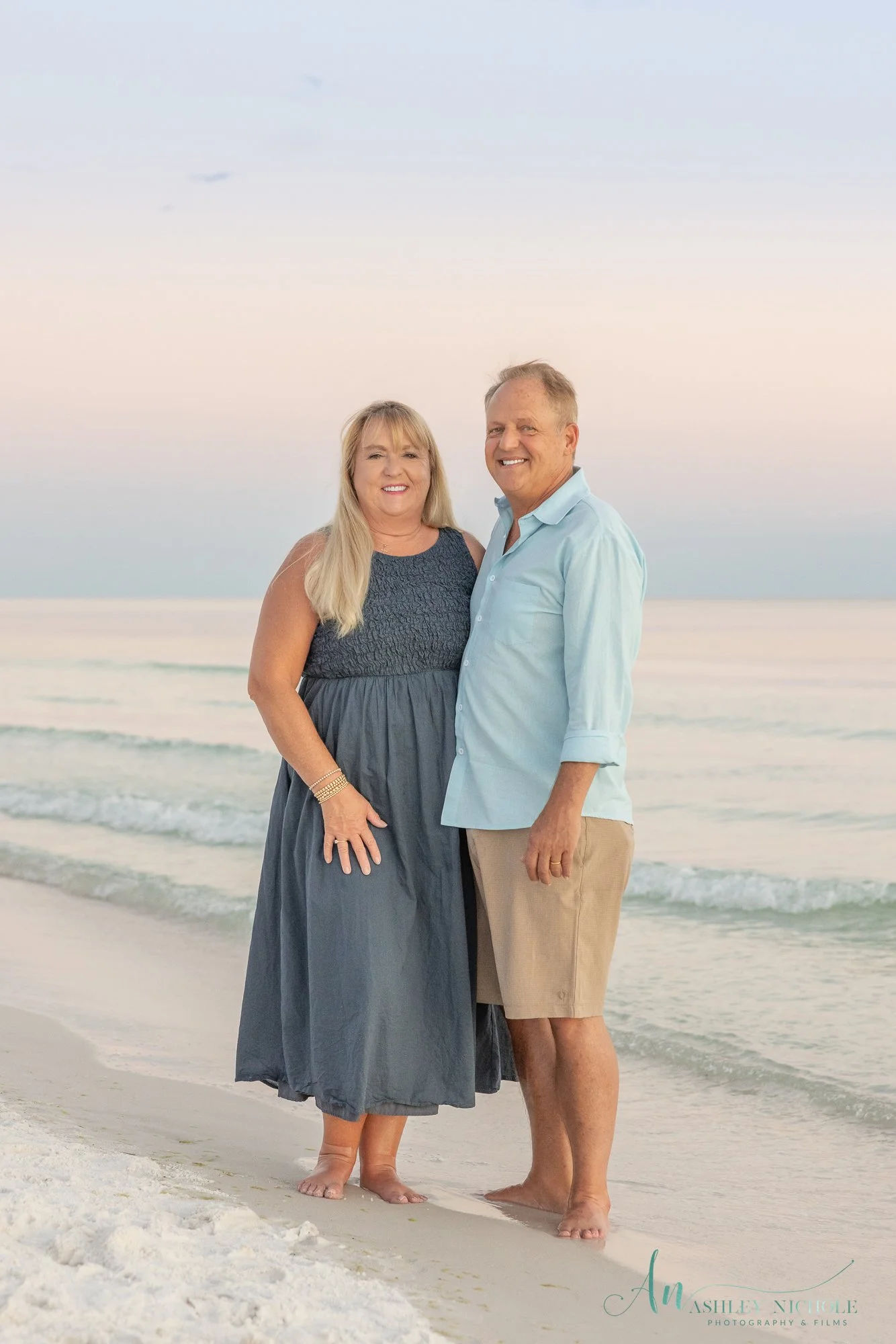 A smiling middle-aged couple standing barefoot on a beach at sunset, with the ocean and sky in the background.