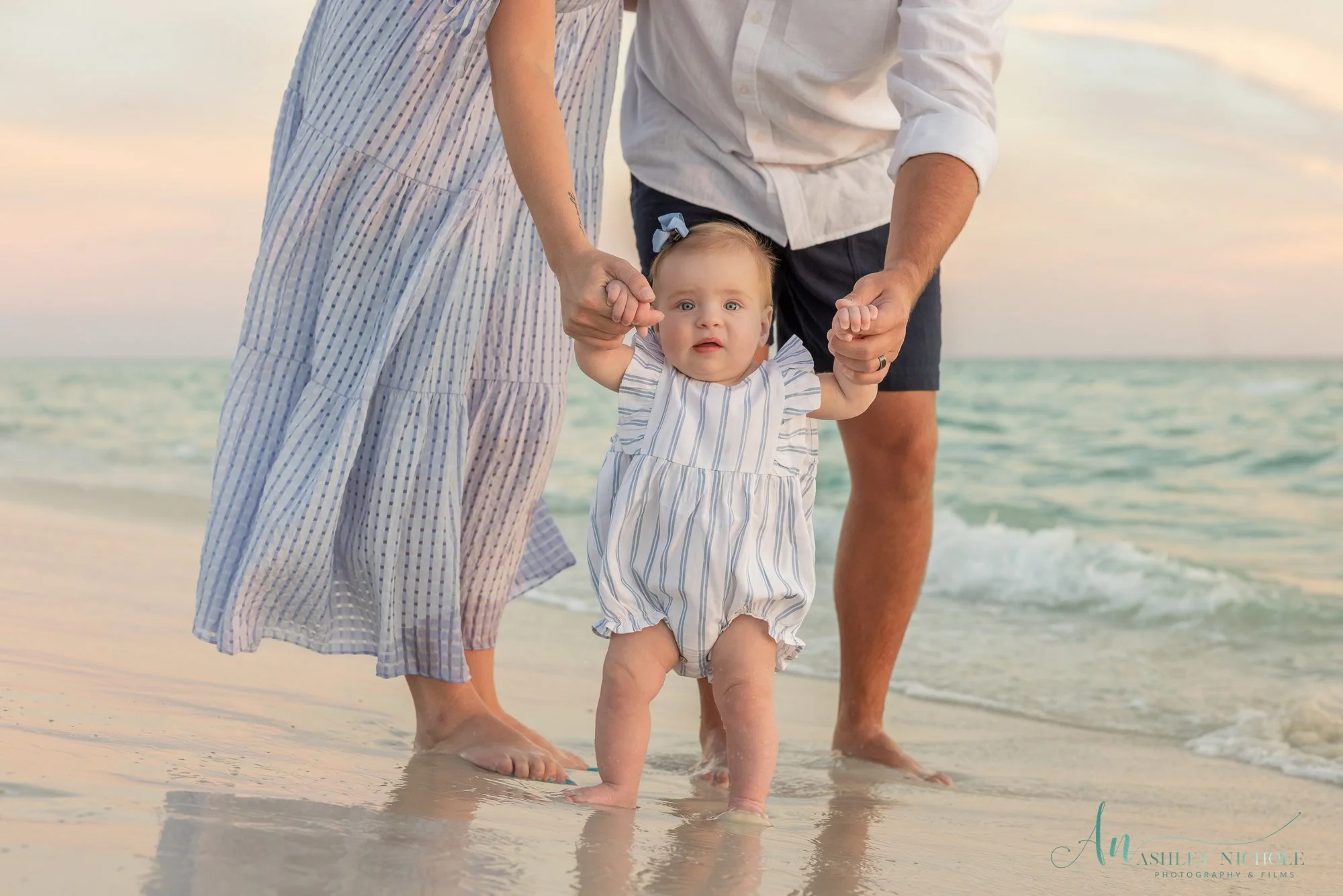 A young child in a striped romper being held by both parents at the beach, with the ocean and sky in the background.