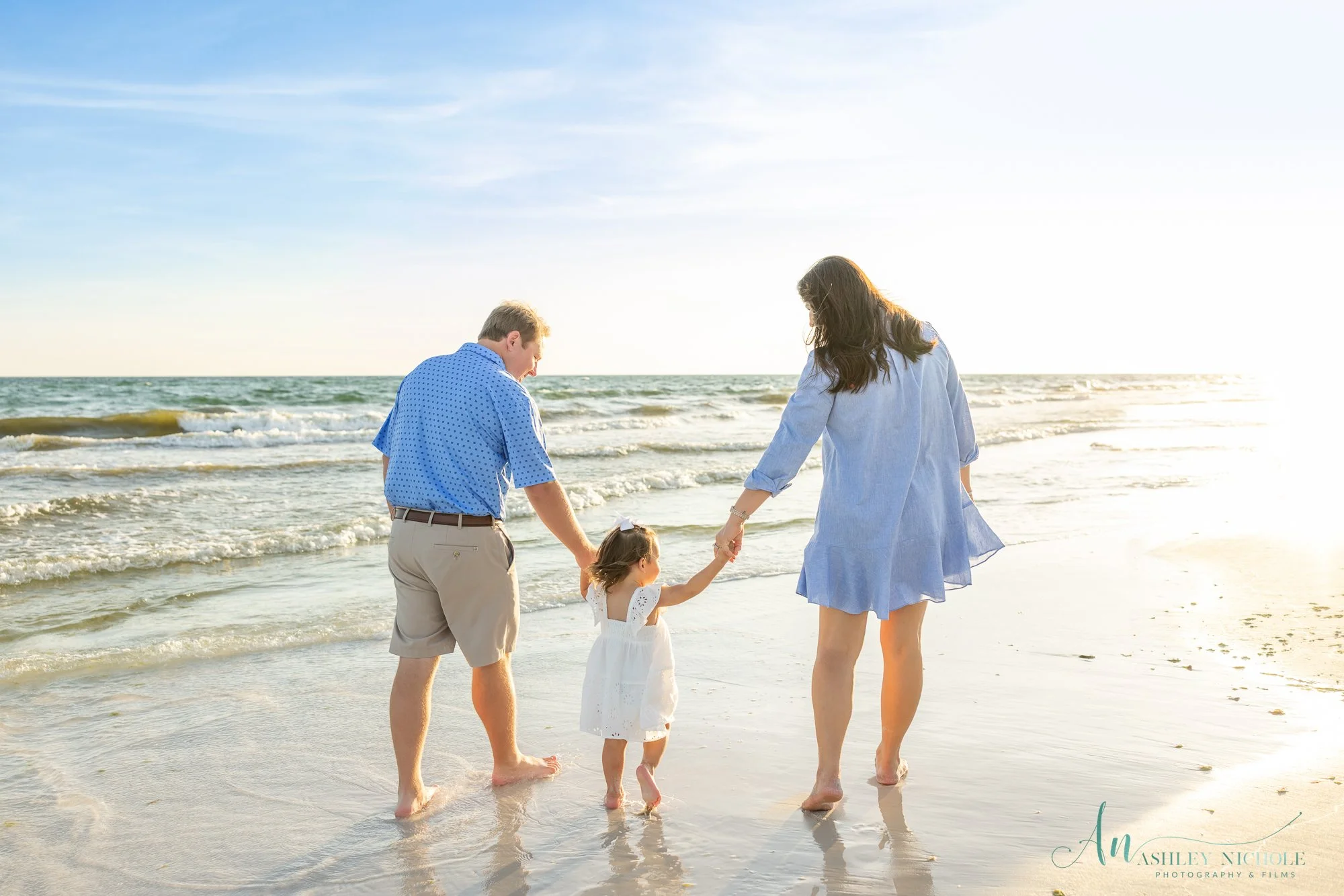 A family of three walking along the beach holding hands, with ocean waves and a clear sky in the background.