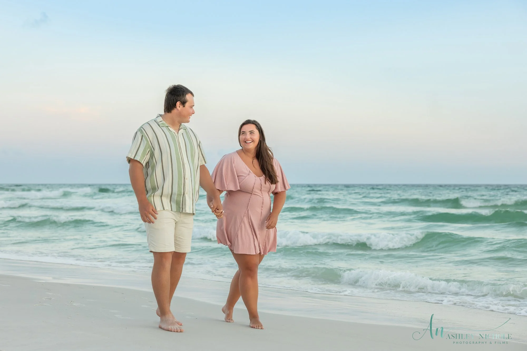 A couple is walking on the beach holding hands, with the ocean waves behind them and a partly cloudy sky above.