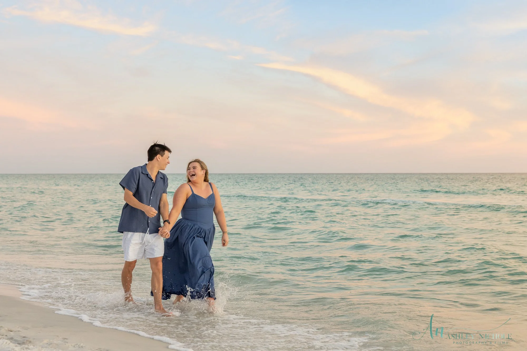 A smiling couple walking hand-in-hand on a sandy beach, near the ocean, during sunset.