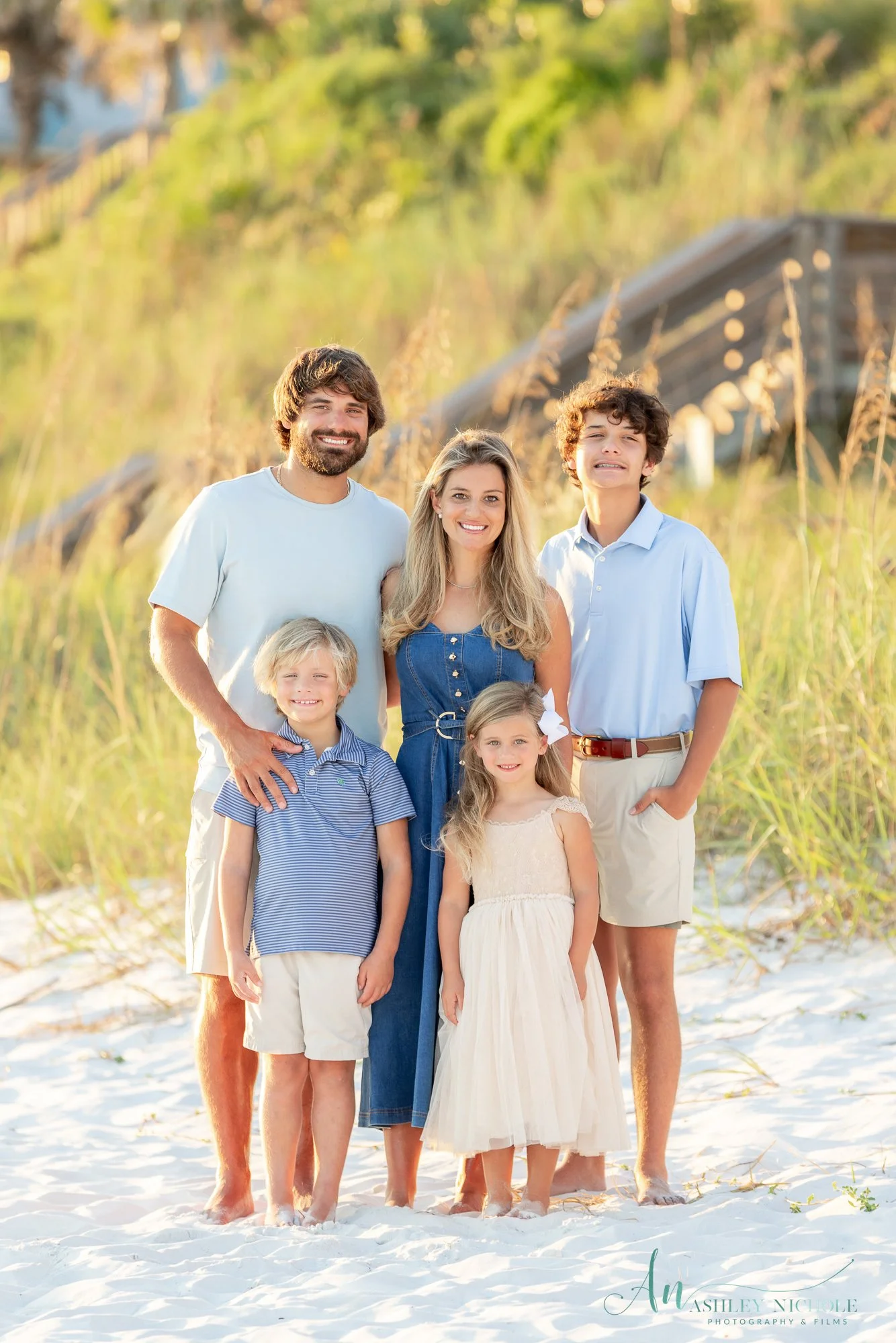 A family of six standing on a sandy beach with tall grass and a wooden structure in the background during sunset, all smiling at the camera.