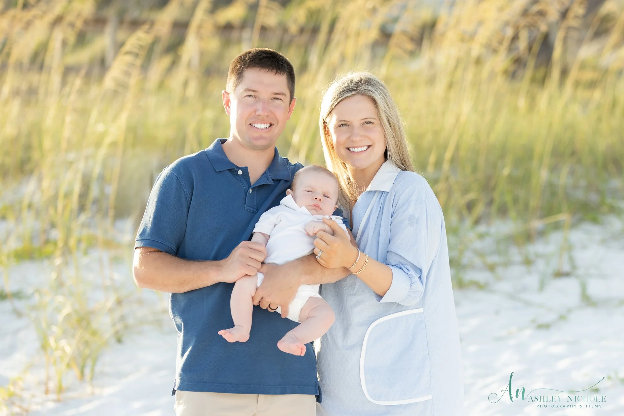 A happy family of three with a baby, outdoors on a sunny day at the beach with tall grass in the background.