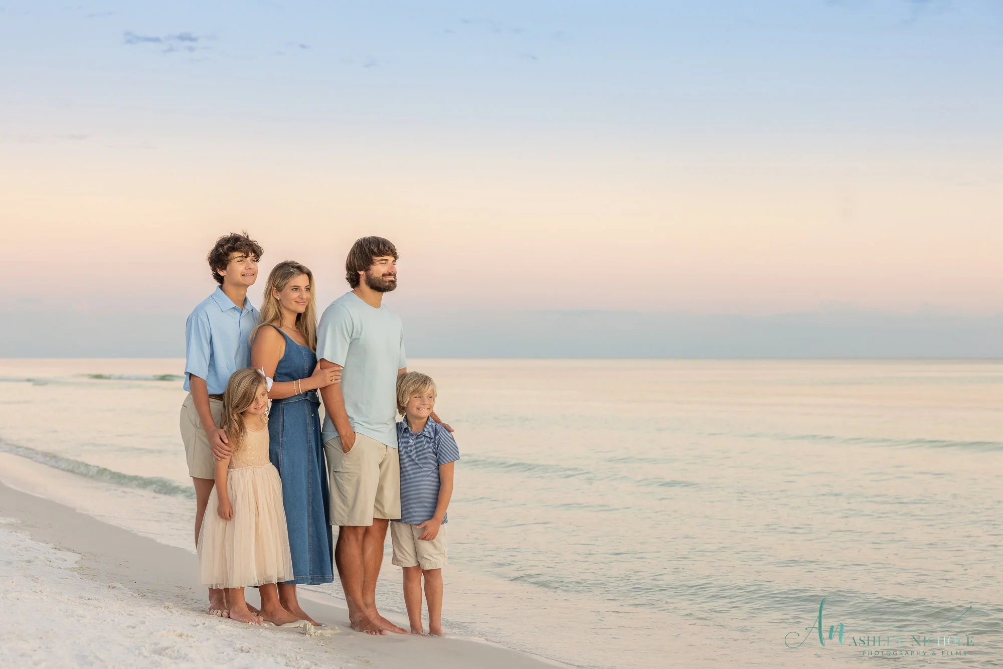 A family of six standing barefoot on the beach at sunset, with the ocean in the background.
