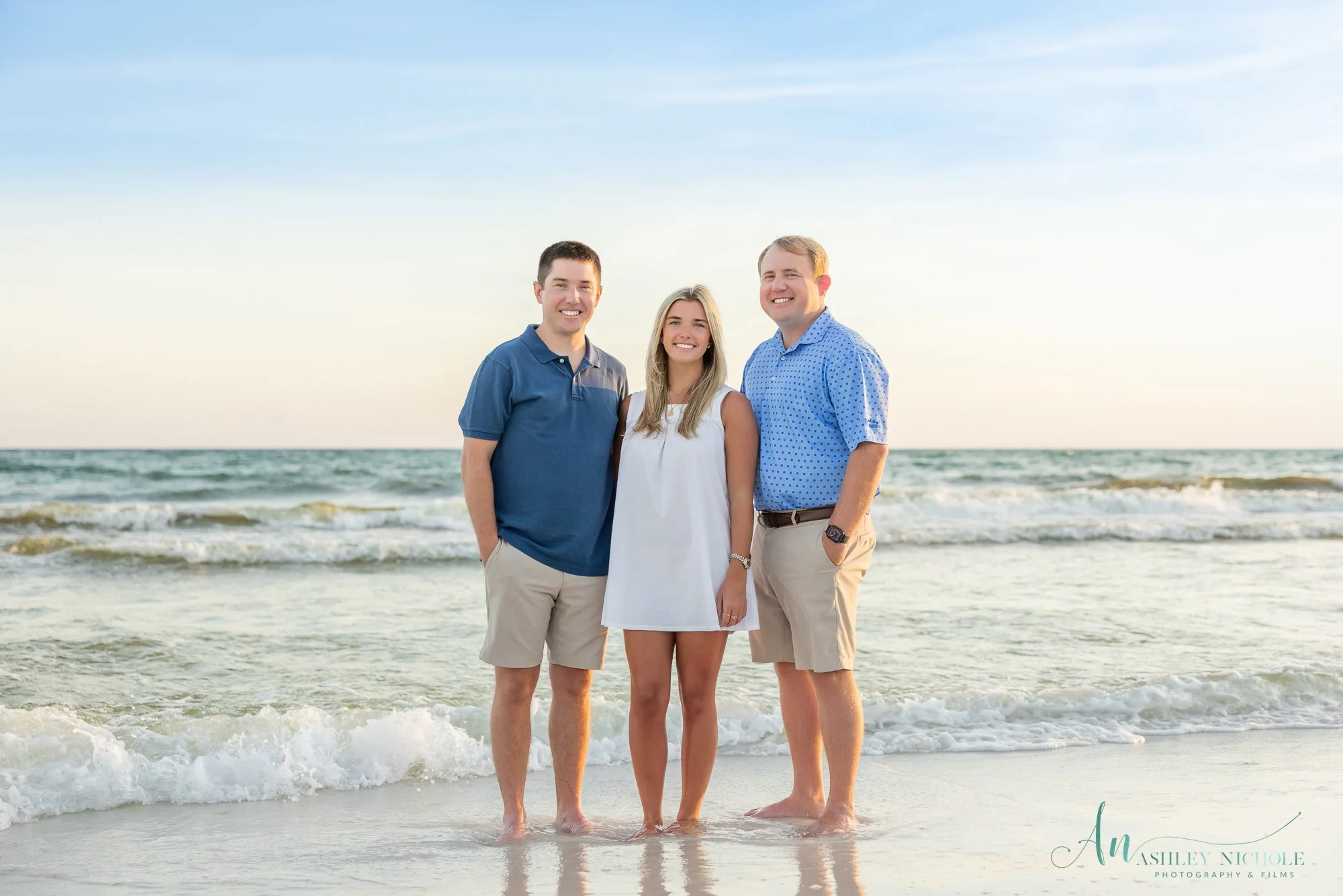 Three young adults, two men and one woman, standing together on the beach with ocean waves in the background, smiling at the camera.